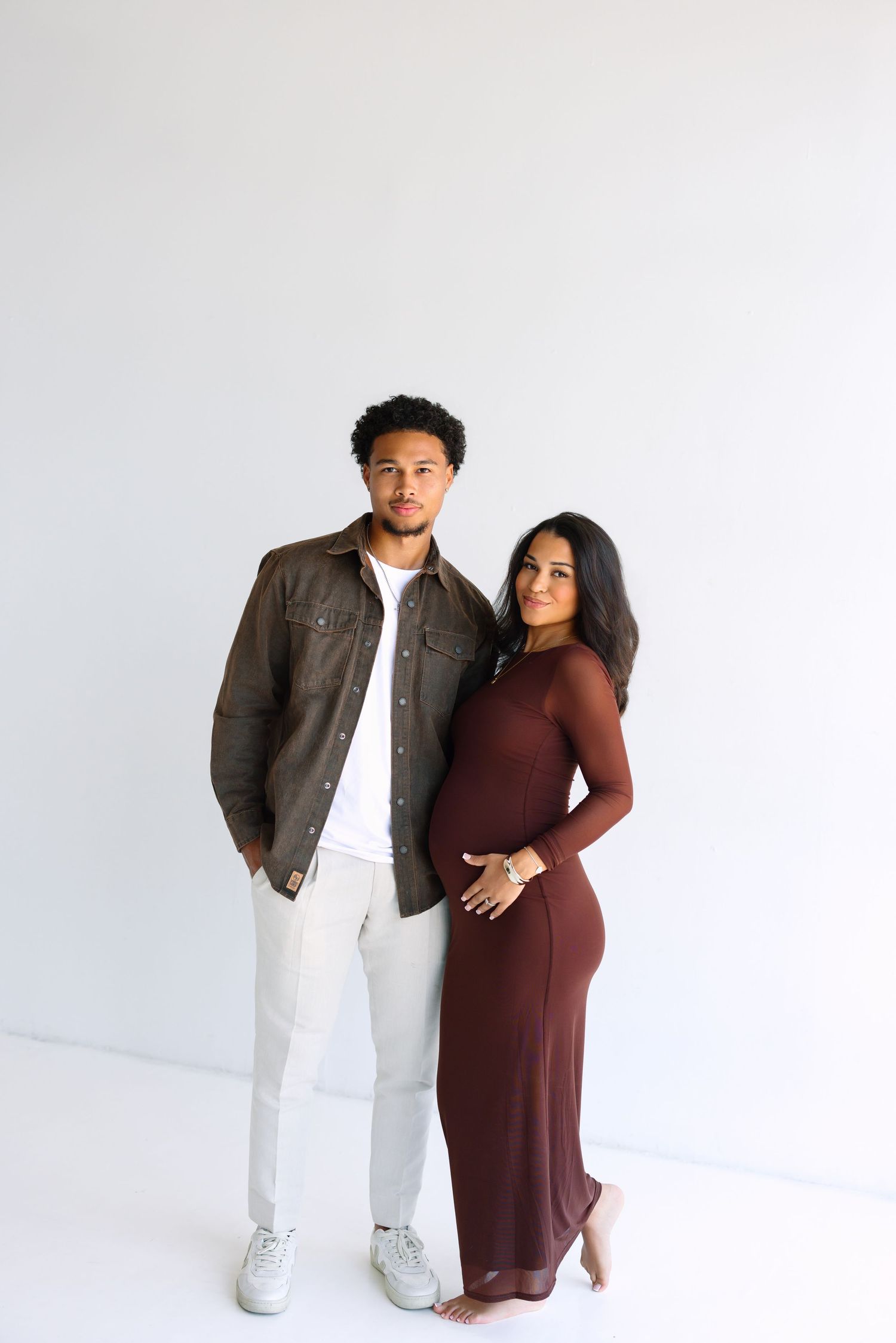 A couple poses together, the woman in a burgundy maxi dress cradling her baby bump, and the man in a brown jacket and white pants.