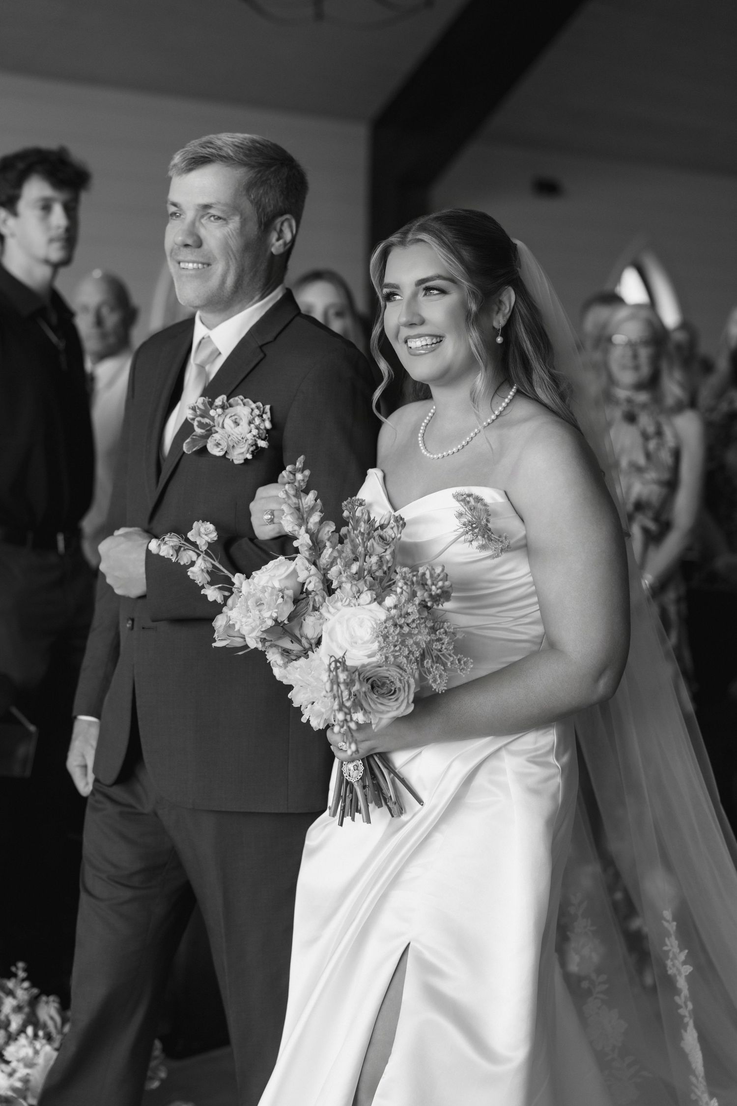 Black and white photo of a smiling bride in strapless gown holding bouquet, walking down the aisle with her father.