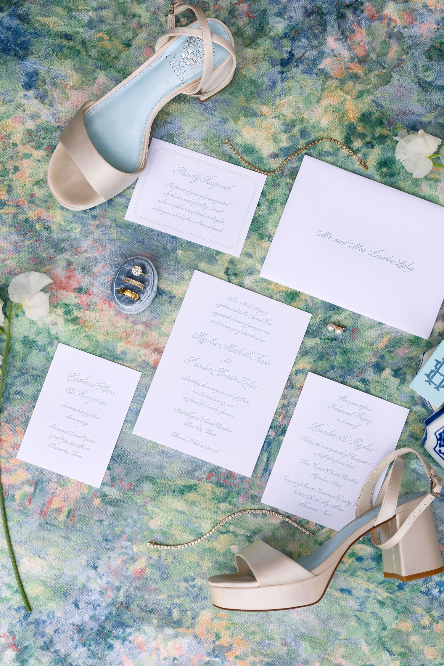 Wedding stationery flat lay with cream heels, rings, and floral accents on watercolor background.