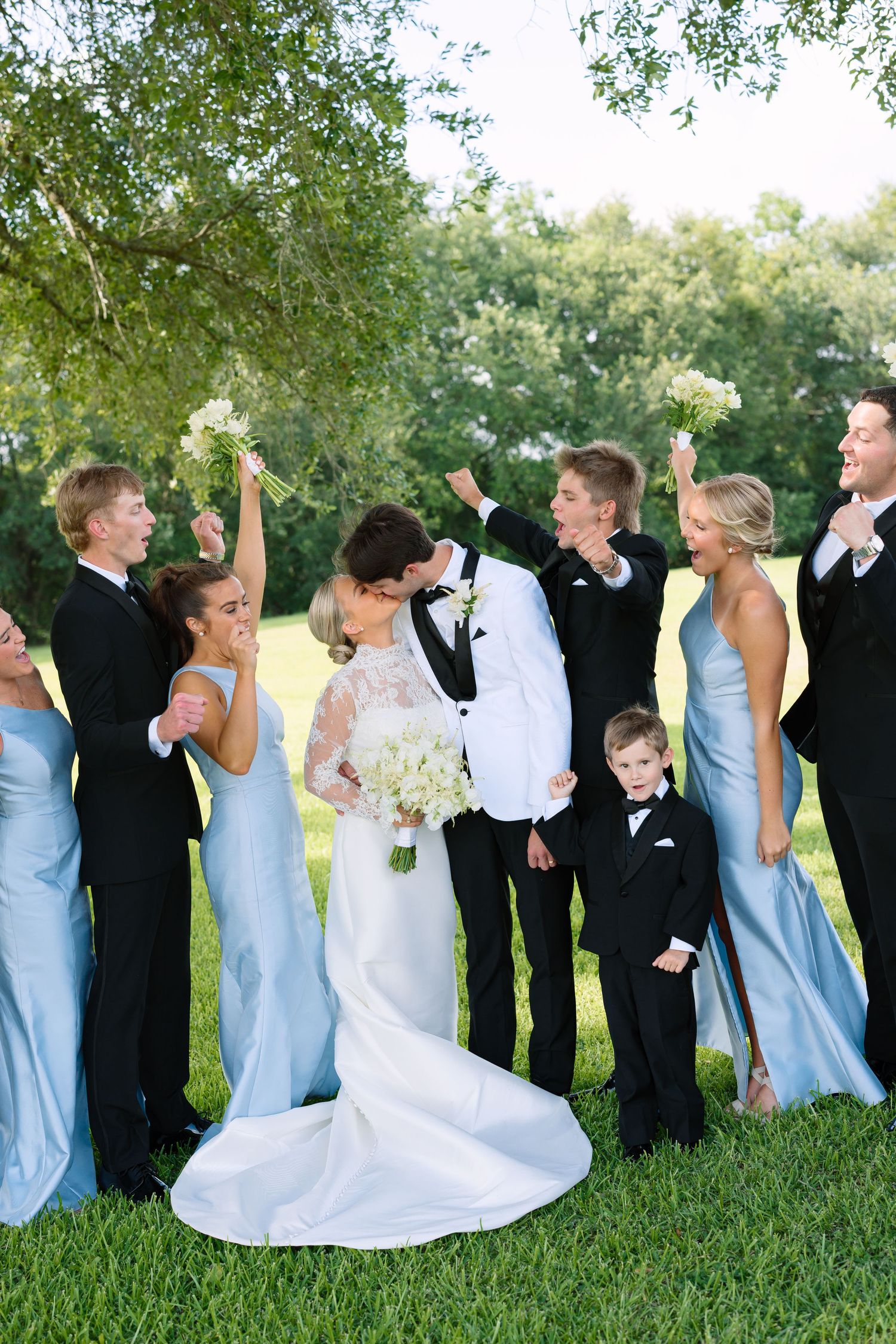 Wedding party celebrating as bride and groom kiss, bridesmaids in blue gowns and groomsmen in black tuxedos outdoors.