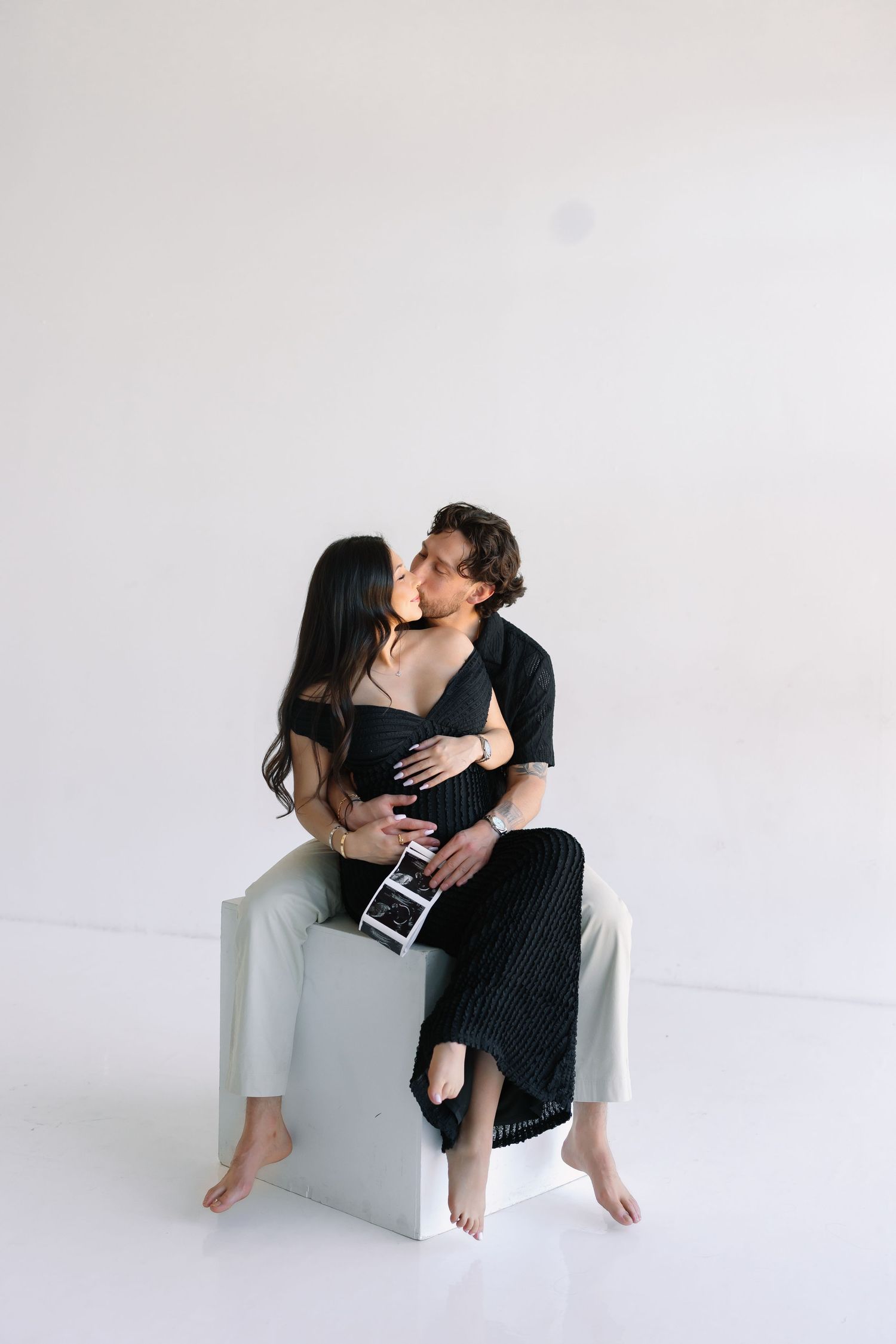 Couple sharing a tender kiss while sitting on a white cube, man embracing pregnant woman holding an ultrasound photo.