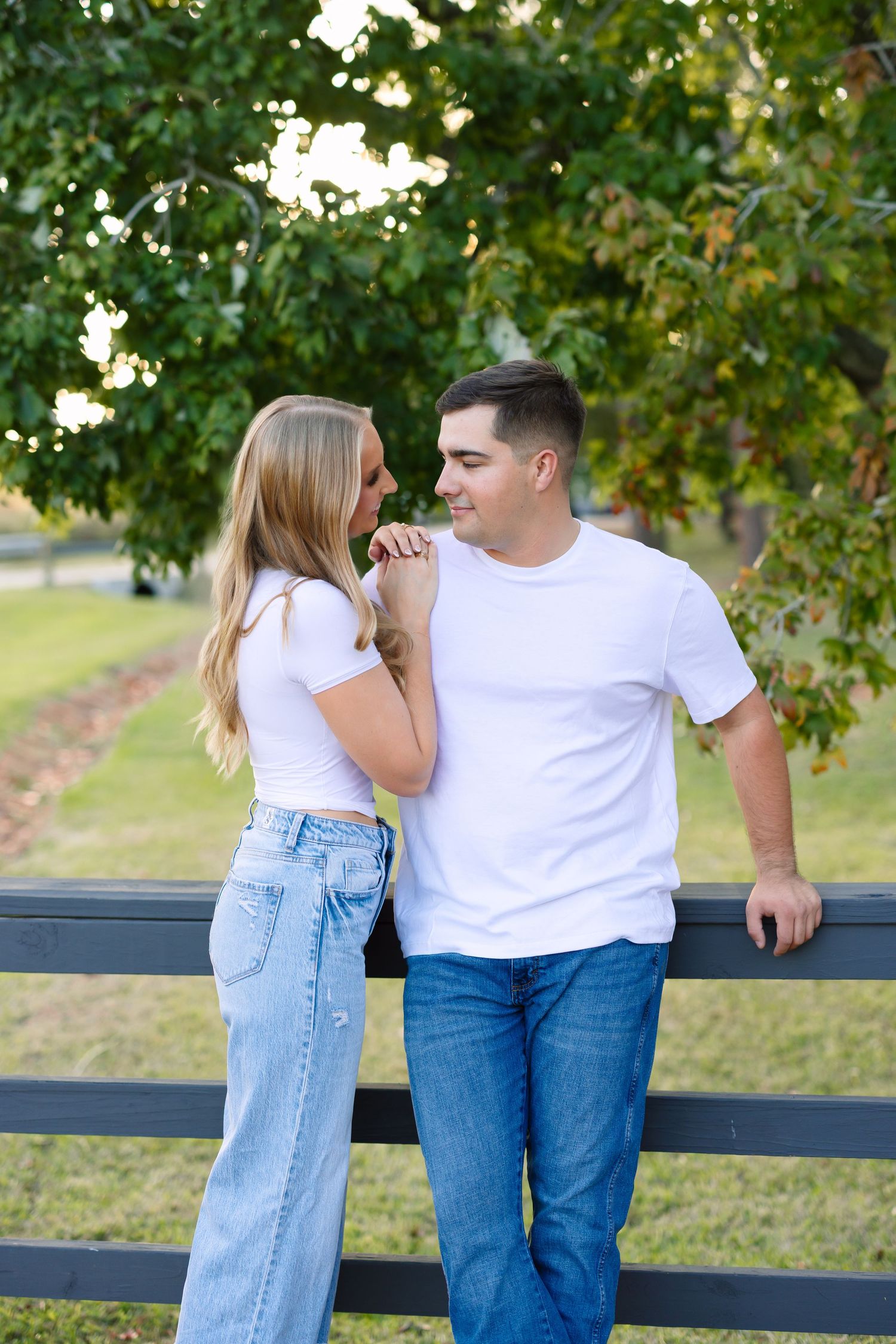 Couple in matching white tops and jeans sharing a tender moment leaning against a black wooden fence outdoors.
