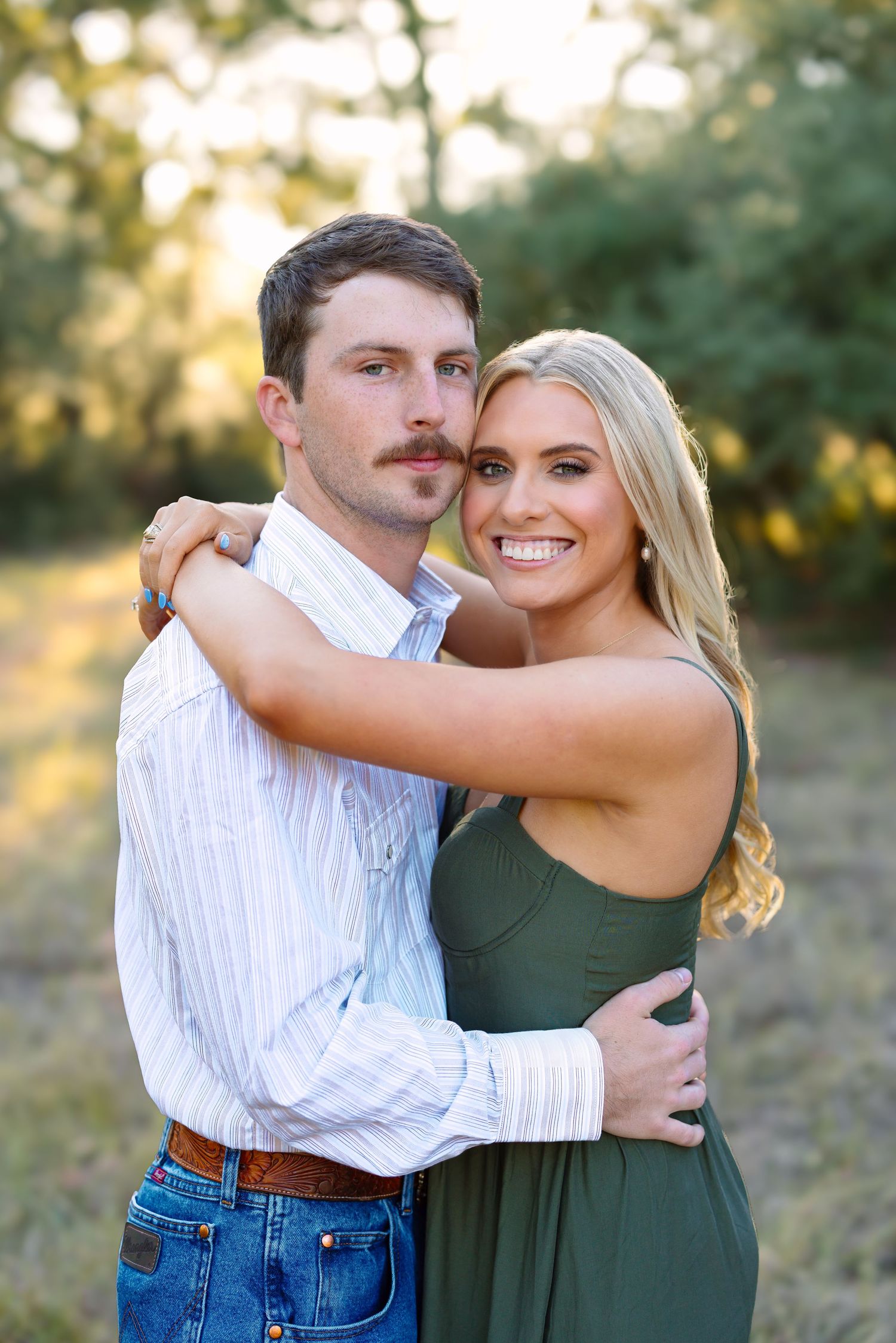 Smiling couple embracing outdoors at golden hour, man in striped shirt and jeans, woman in olive green dress.