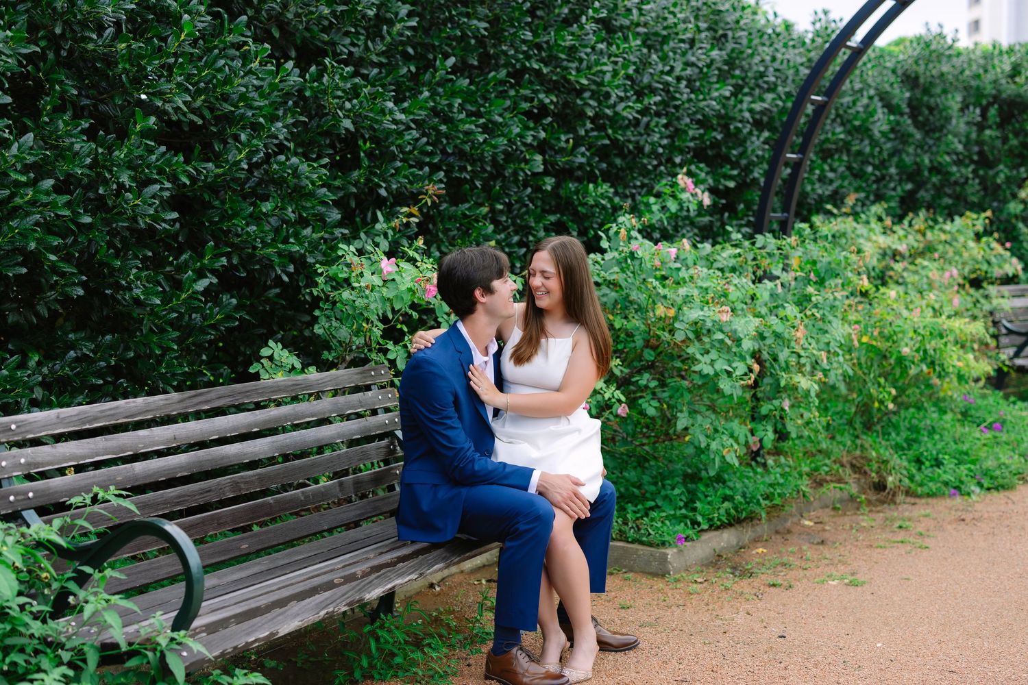 A couple embraces on a garden path surrounded by green foliage and wooden benches.