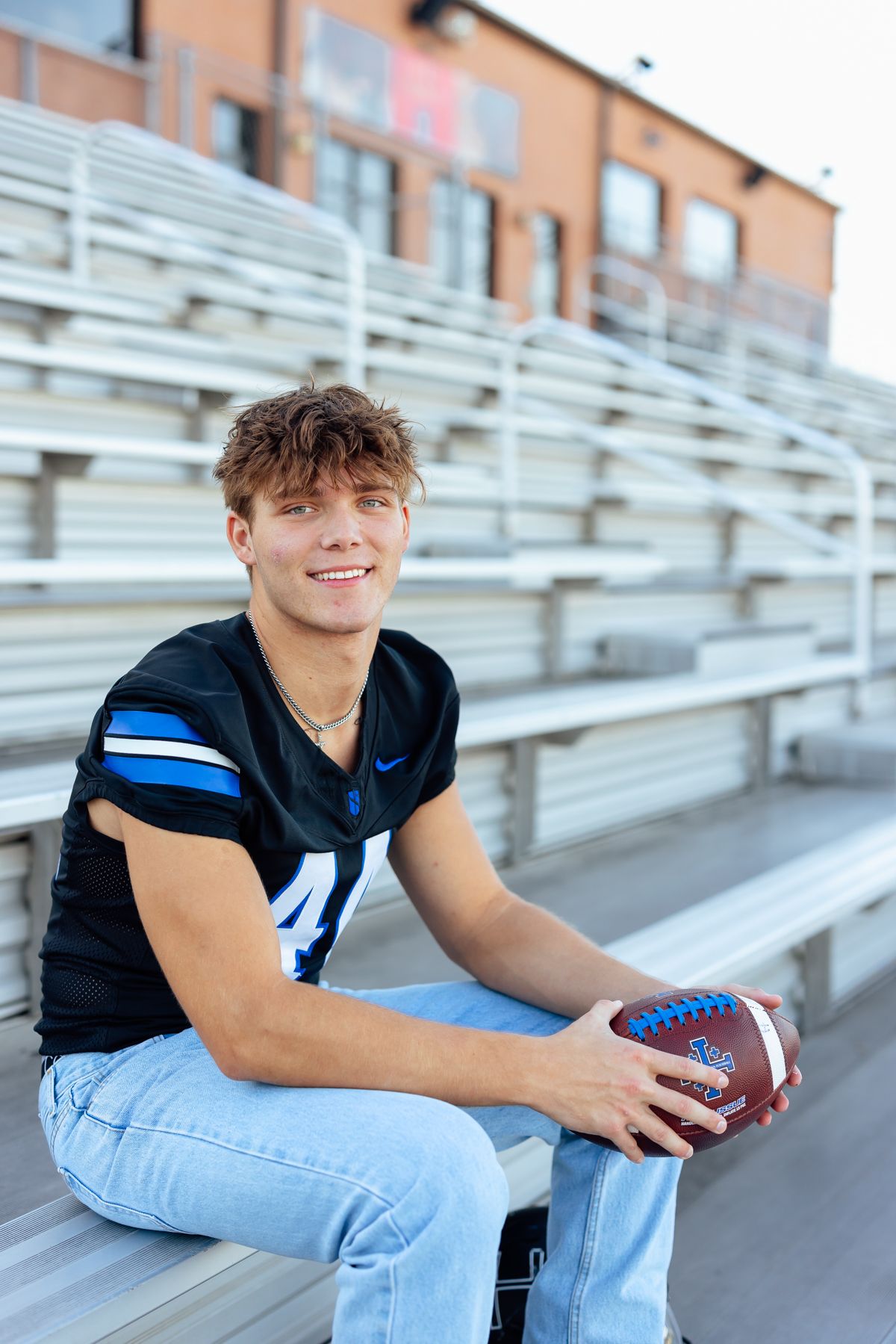 Football player in black jersey and light jeans sits on bleachers holding a football at a stadium.