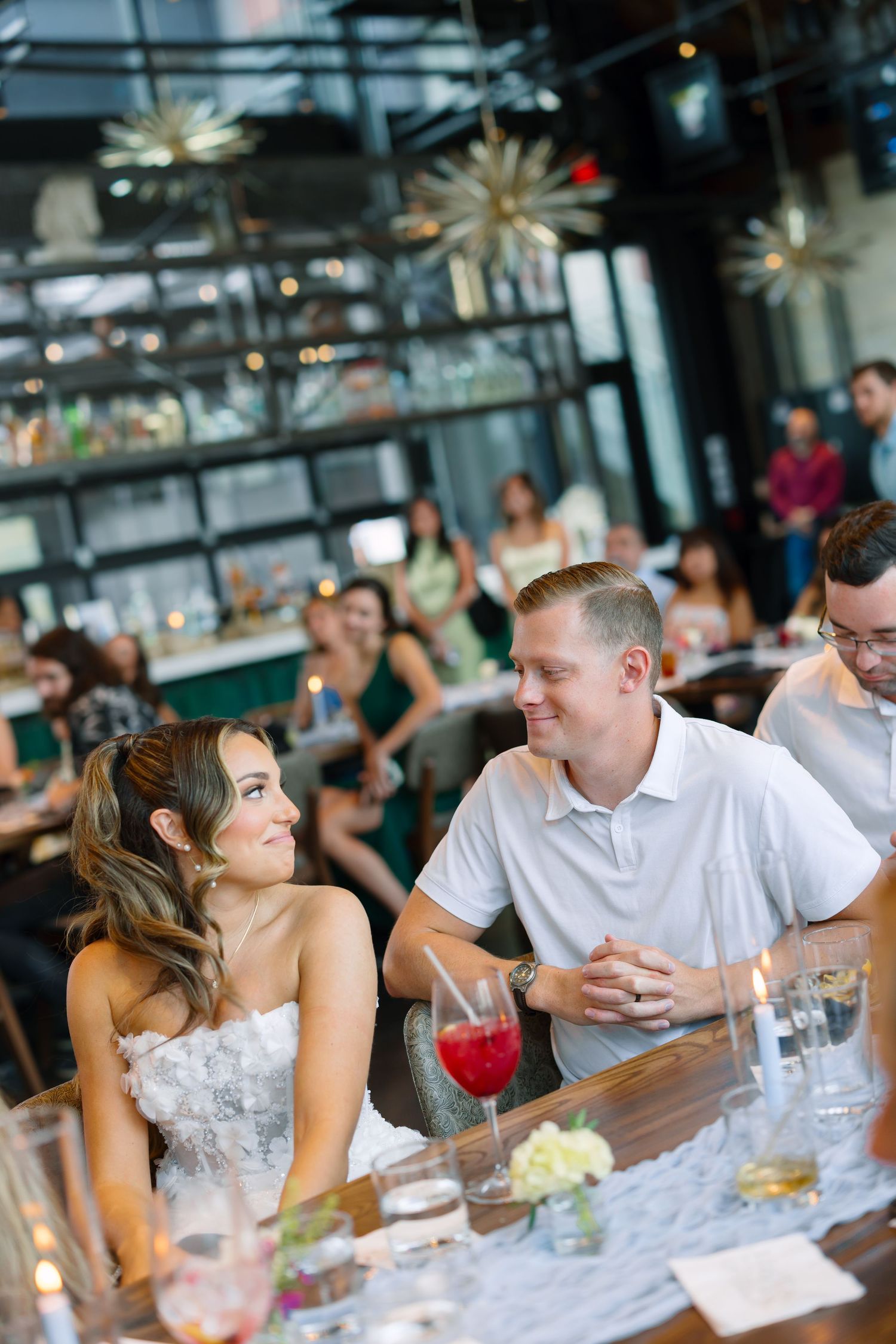 Bride and groom share a joyful moment at their wedding reception table, surrounded by guests in a modern venue.