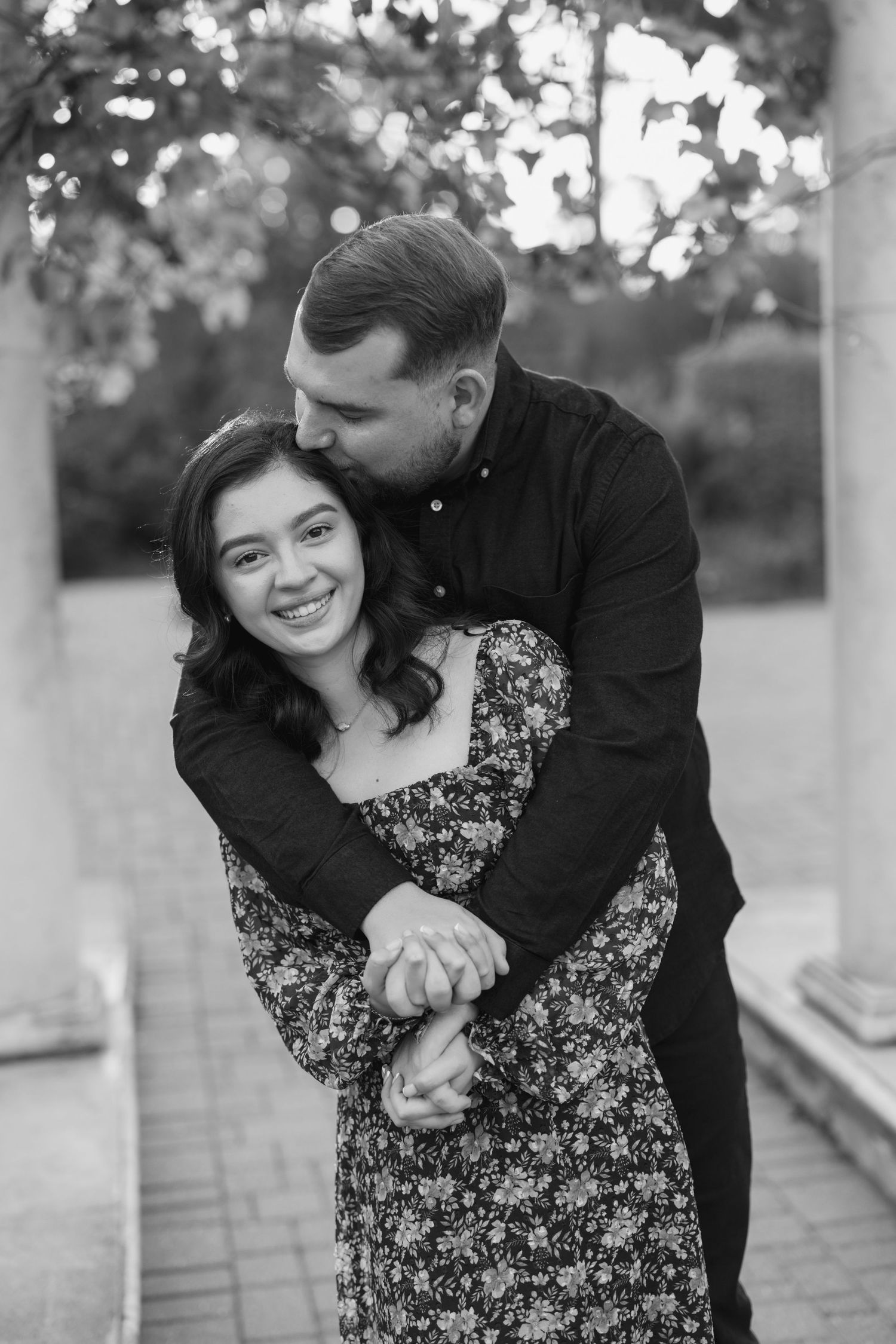 Black and white photo of a couple embracing outdoors, man kissing woman's head while she smiles.