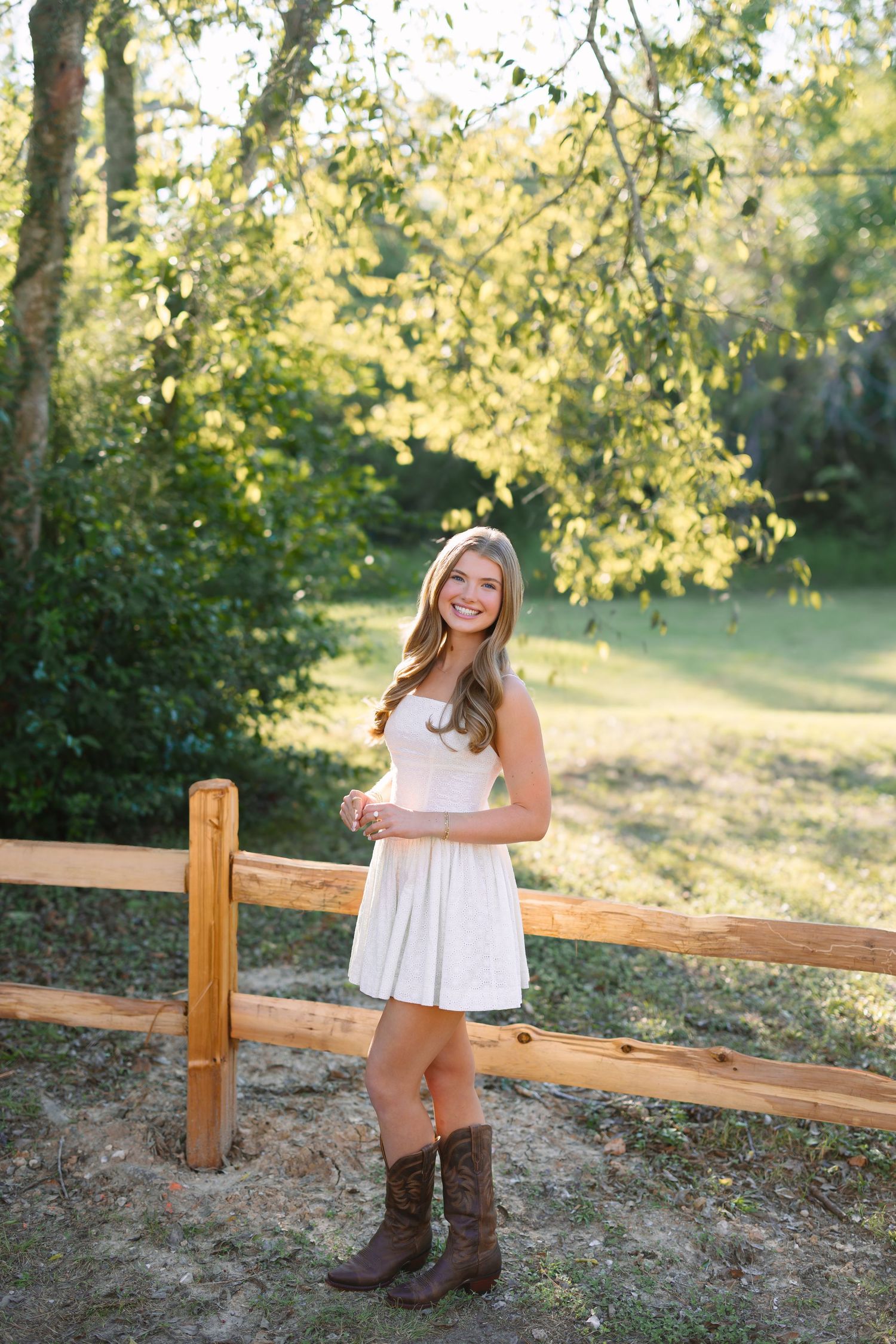 Young woman in white dress and cowboy boots smiling by a wooden fence in a sunny green outdoor setting.