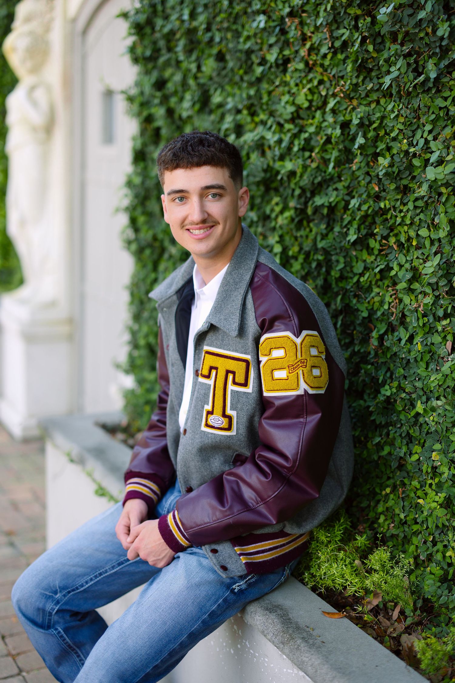 Smiling teen in a gray and maroon varsity letterman jacket with T and 26 patches, sitting outdoors by ivy wall.