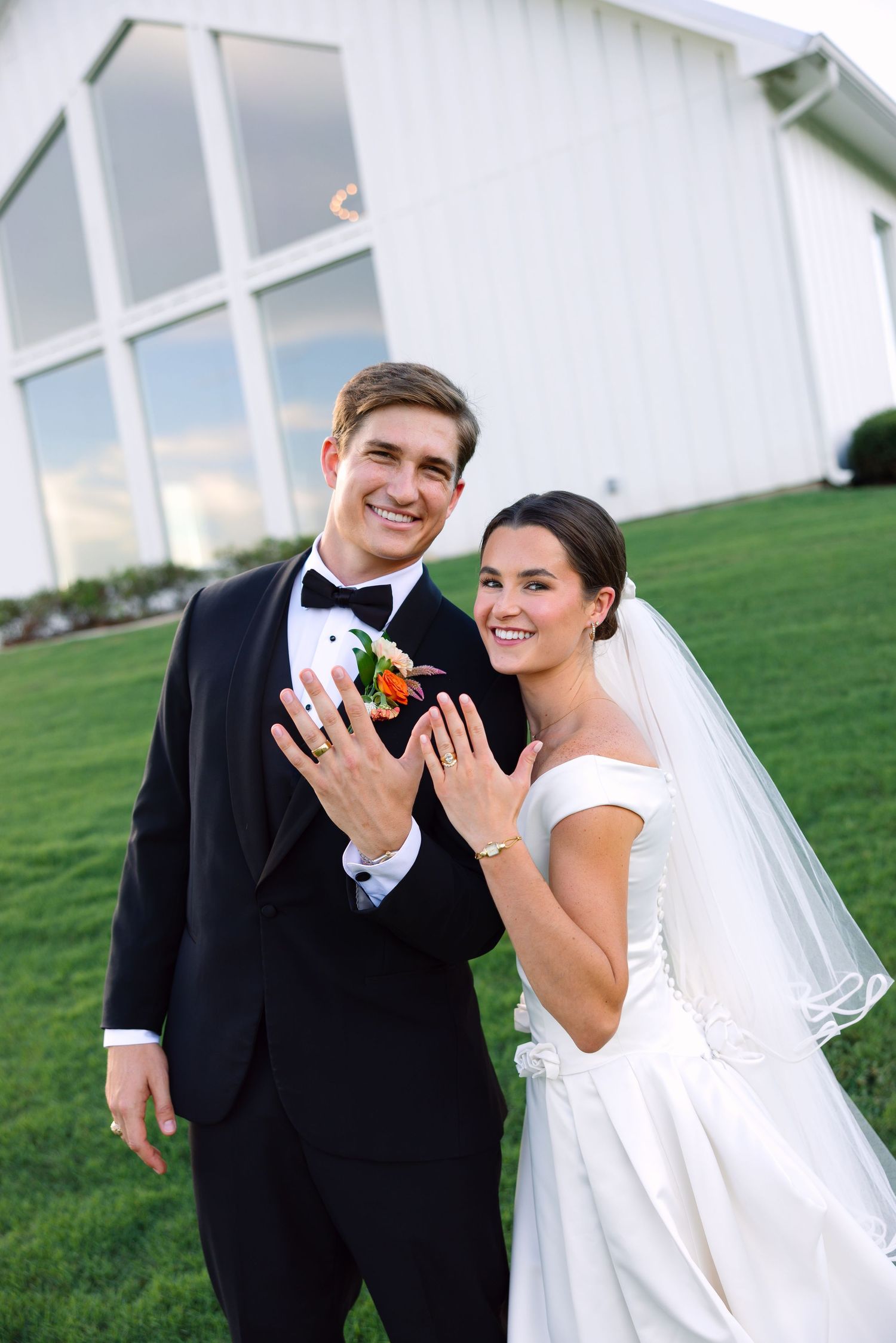 Smiling bride and groom showing wedding rings outside white barn venue, bride in white gown with veil, groom in black tuxedo.