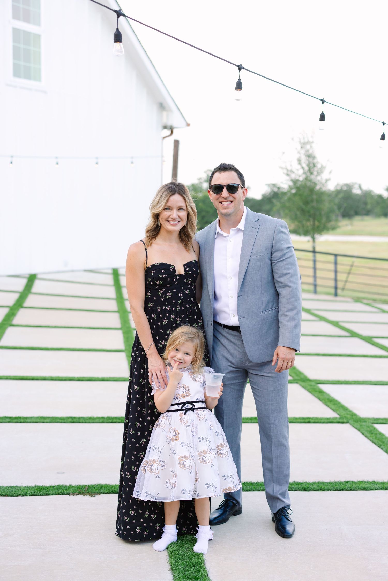 Family posing together in formal attire against a modern white barn backdrop with string lights.