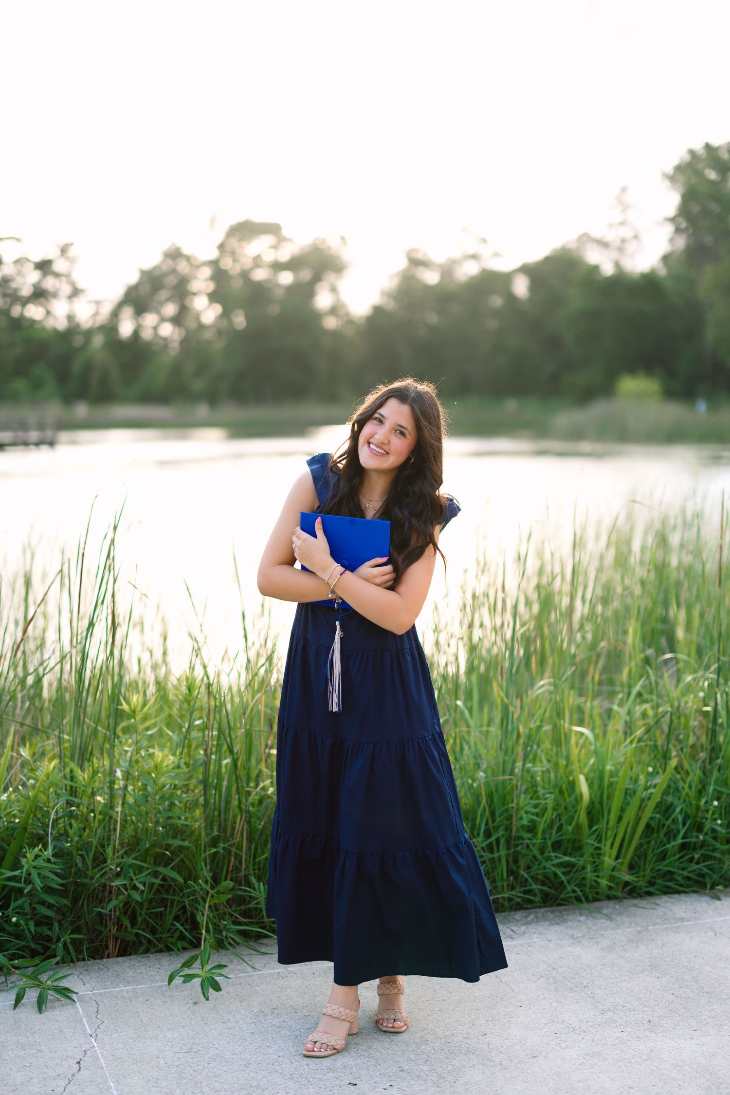 A person in a flowing navy dress poses gracefully by a lakeside with tall reeds.