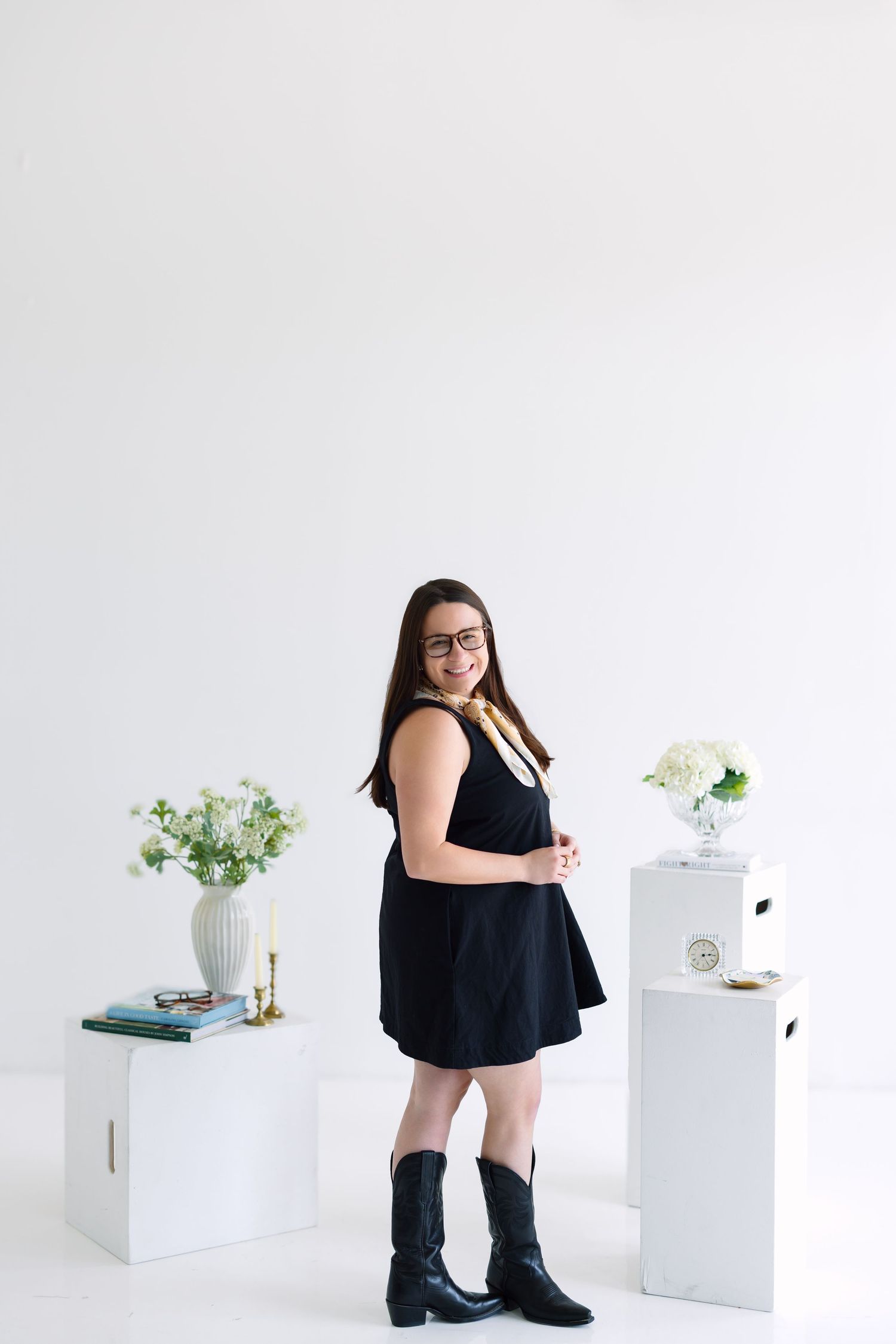 Woman in black dress and cowboy boots poses in white studio with floral arrangements on white pedestals.