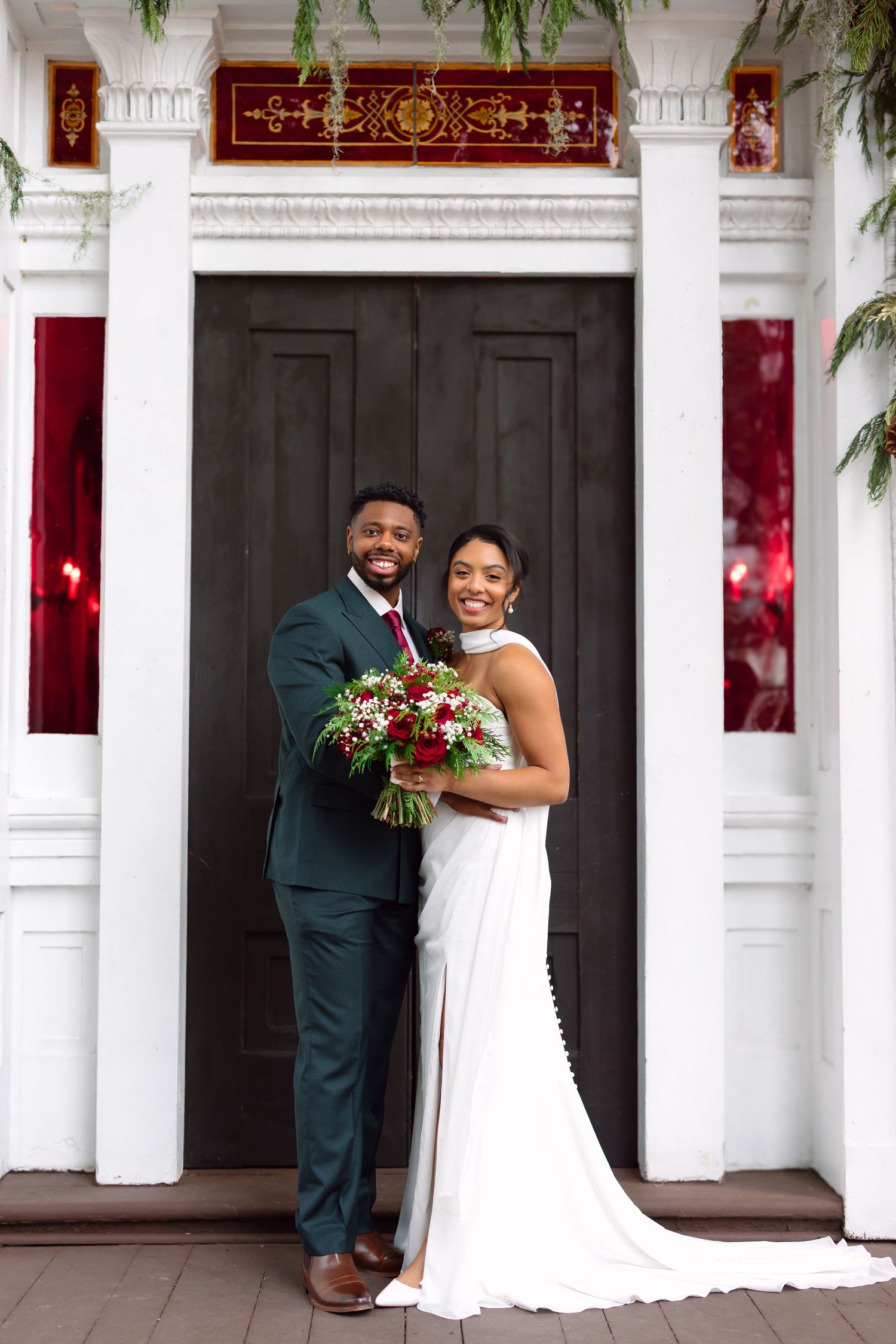 Smiling bride and groom in wedding attire pose before elegant dark doors with white columns and red decor.