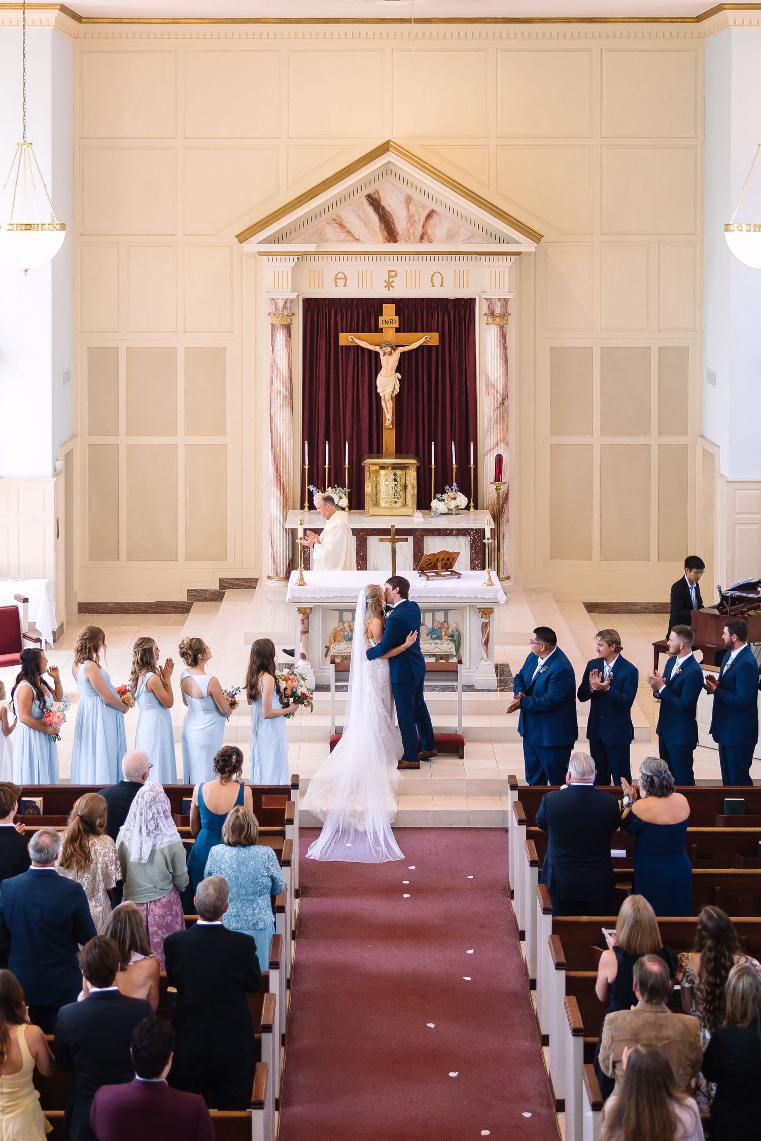 Bride and groom exchange vows at a Catholic church altar surrounded by bridesmaids in blue and groomsmen in navy suits.
