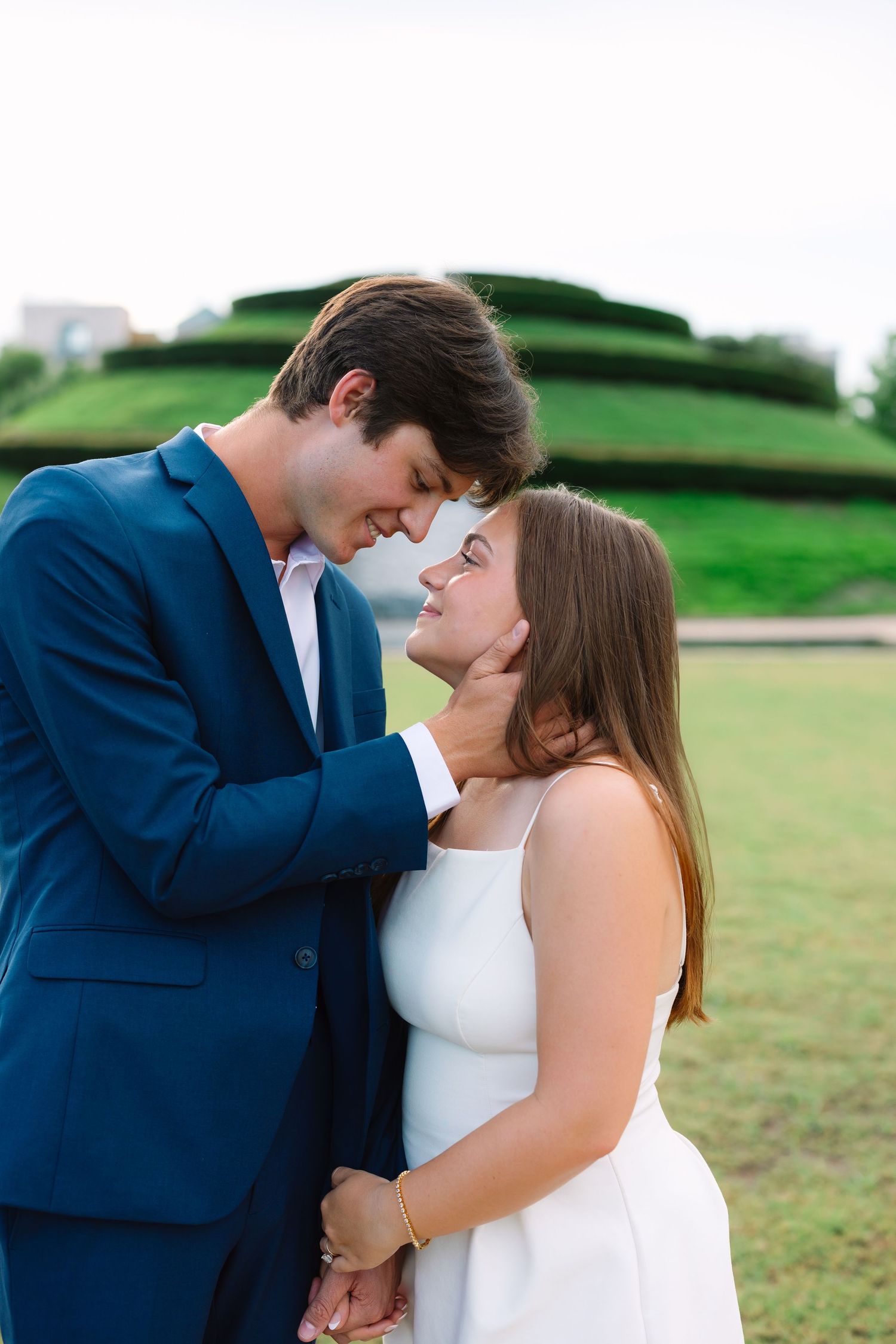 A romantic couple shares an intimate moment on a grassy hill, with the person in navy suit embracing their partner in white.