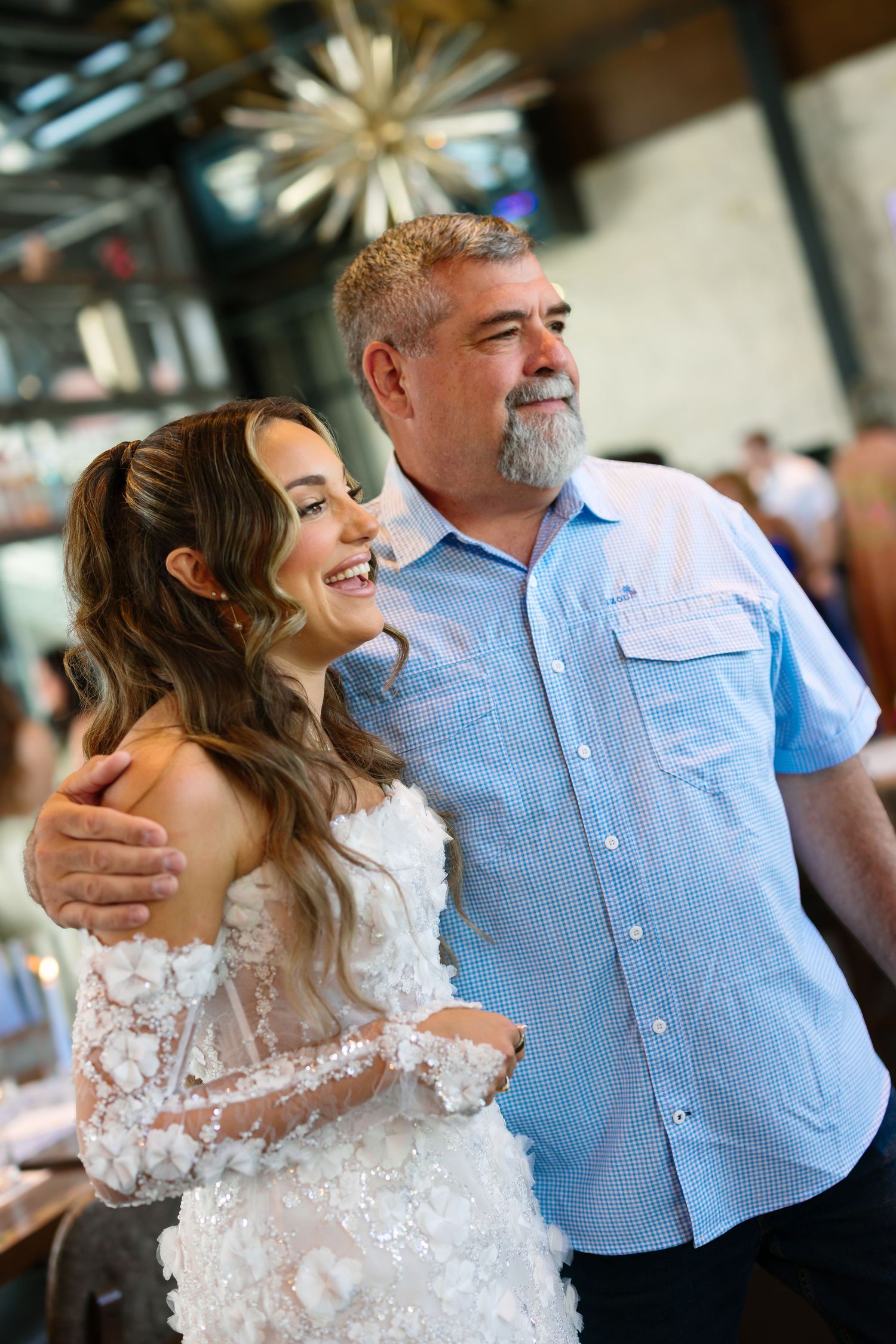 Bride in white lace dress smiling with older man in blue shirt at wedding reception venue.