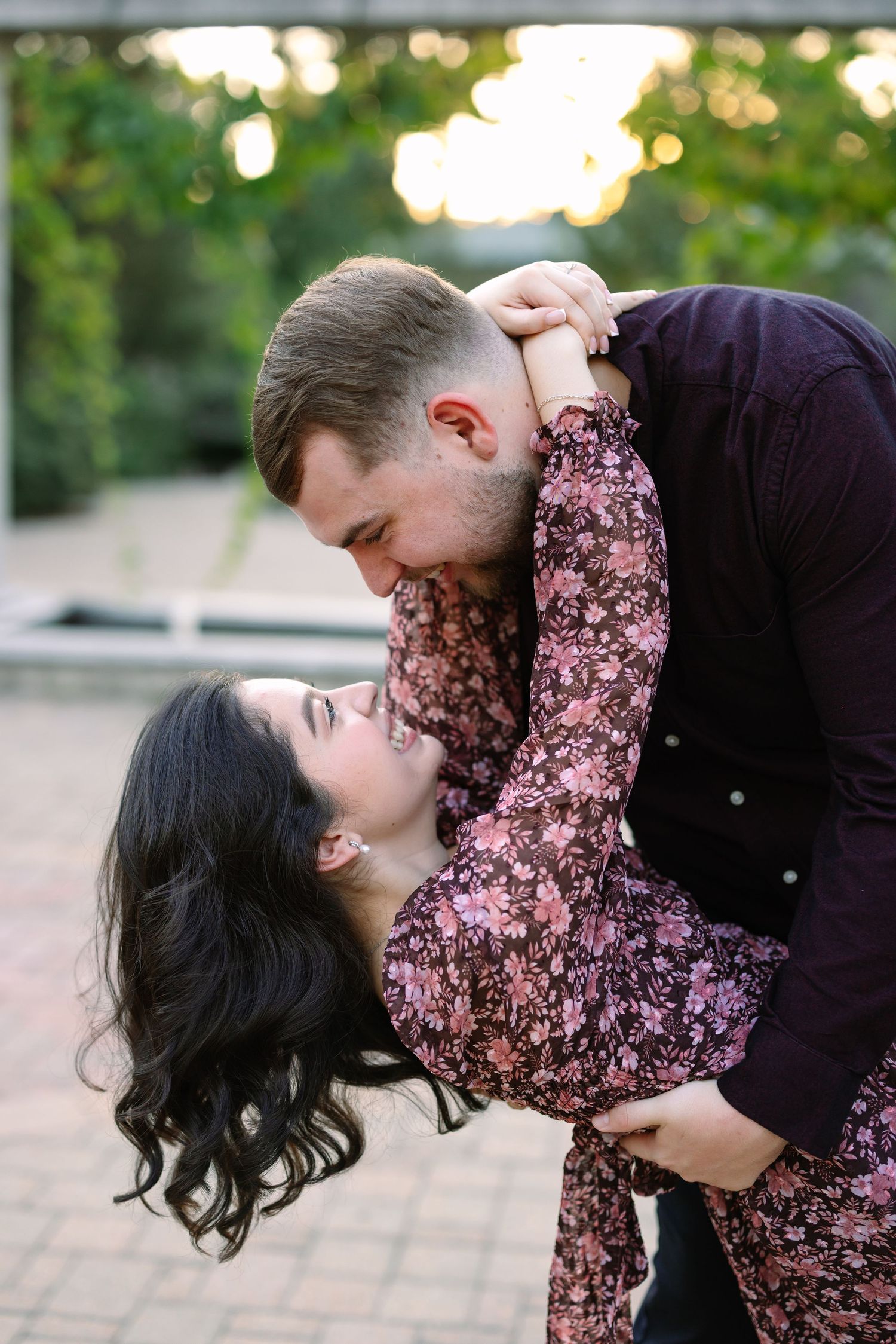 Couple sharing a romantic dip kiss outdoors, woman in floral dress laughing as man in burgundy shirt holds her.