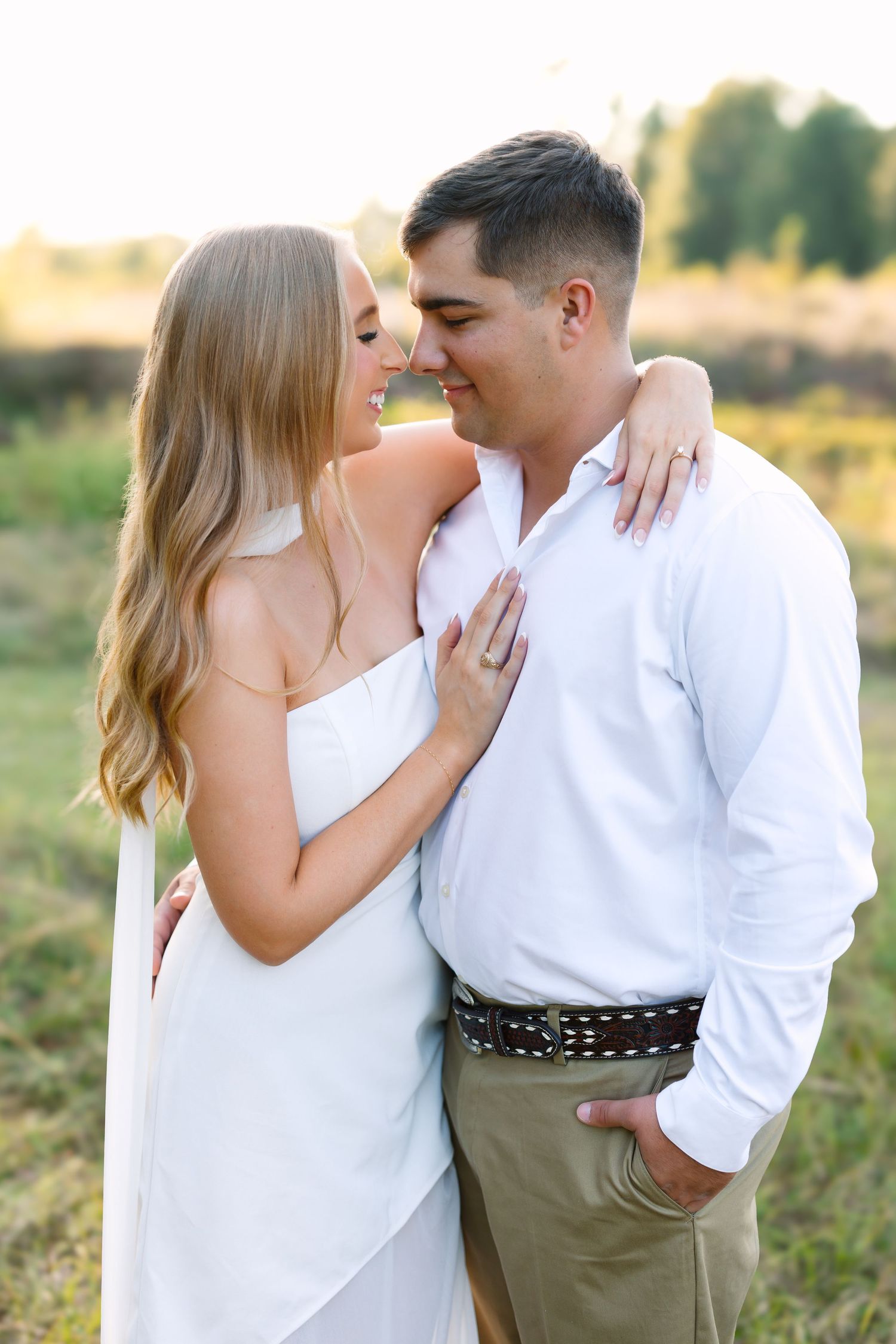 Couple in white outfits sharing a romantic moment outdoors in a sunlit field during golden hour.