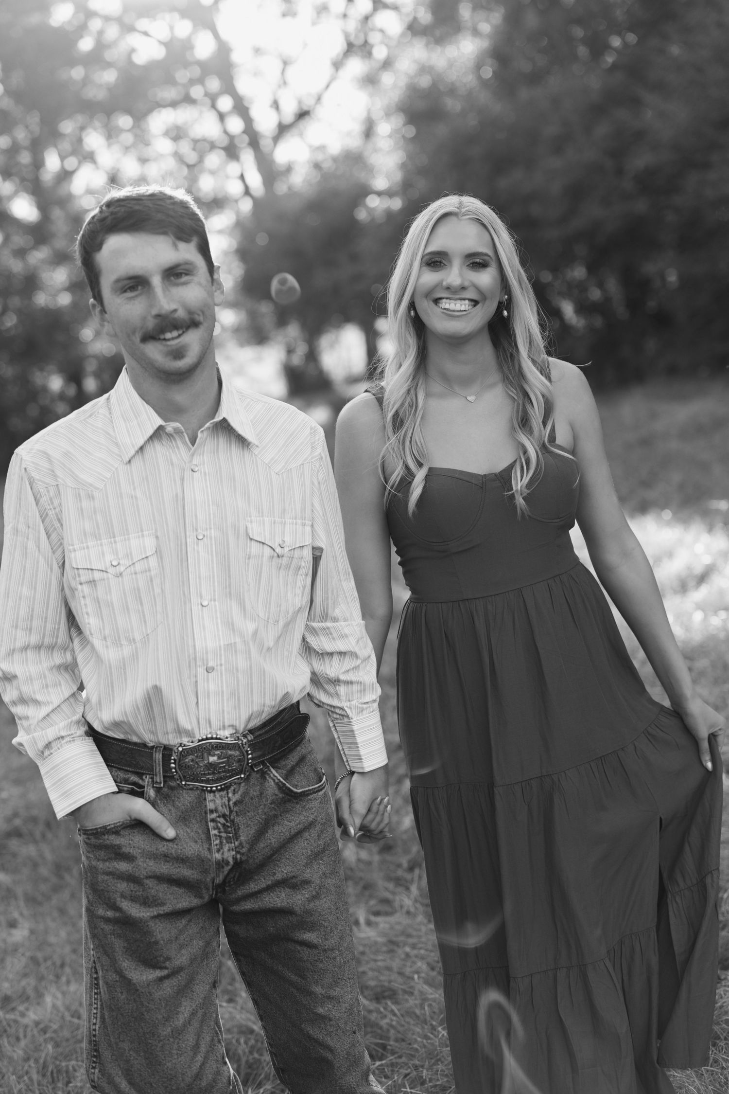 Black and white photo of a smiling couple holding hands outdoors, man in striped shirt and jeans, woman in maxi dress.