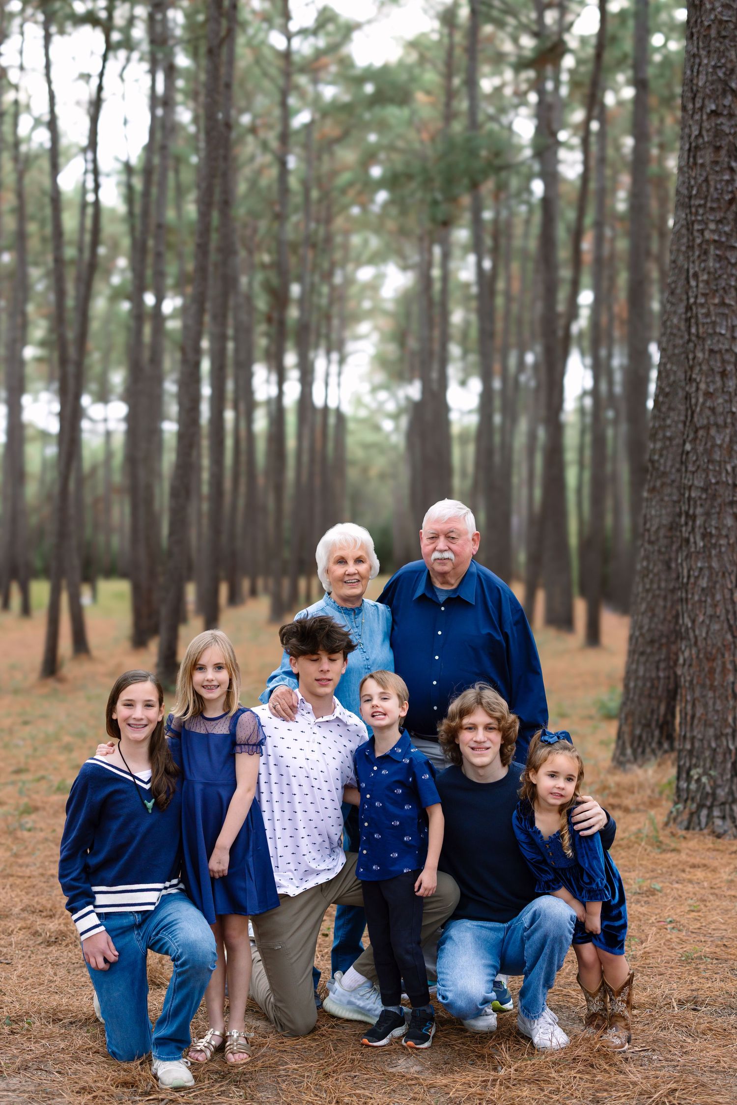 Multi-generational family portrait with grandparents and five grandchildren in a pine forest.