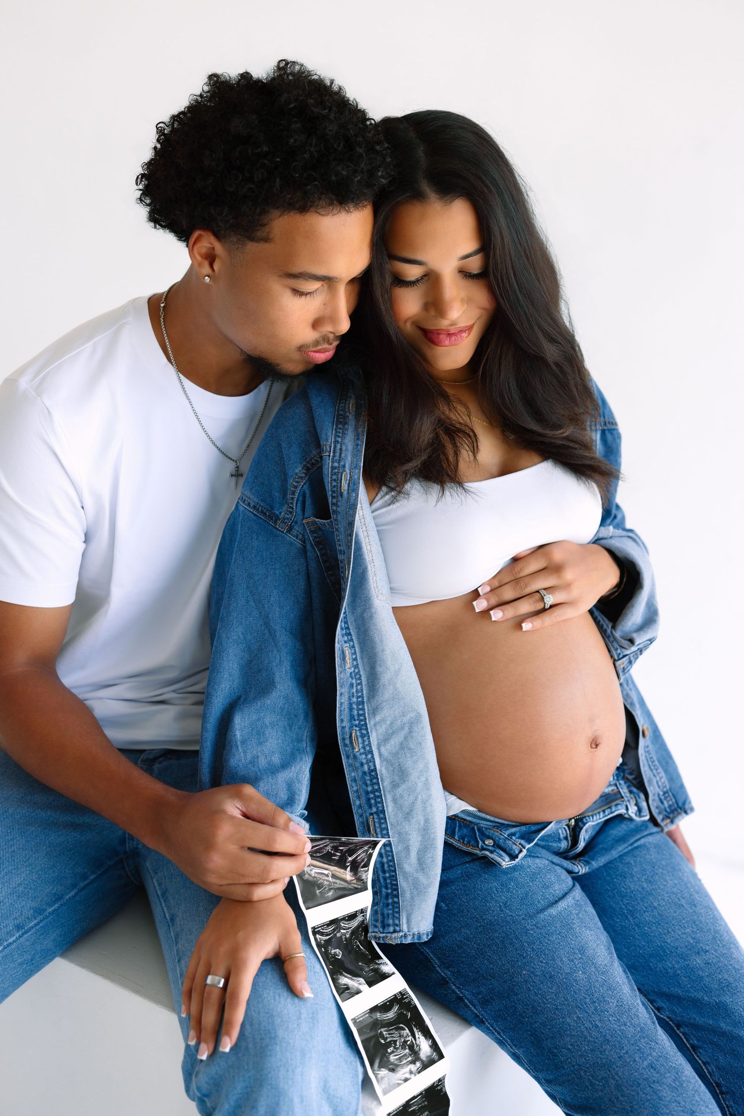 Happy couple in denim outfits holding ultrasound photos, pregnant woman cradling baby bump during maternity photoshoot.