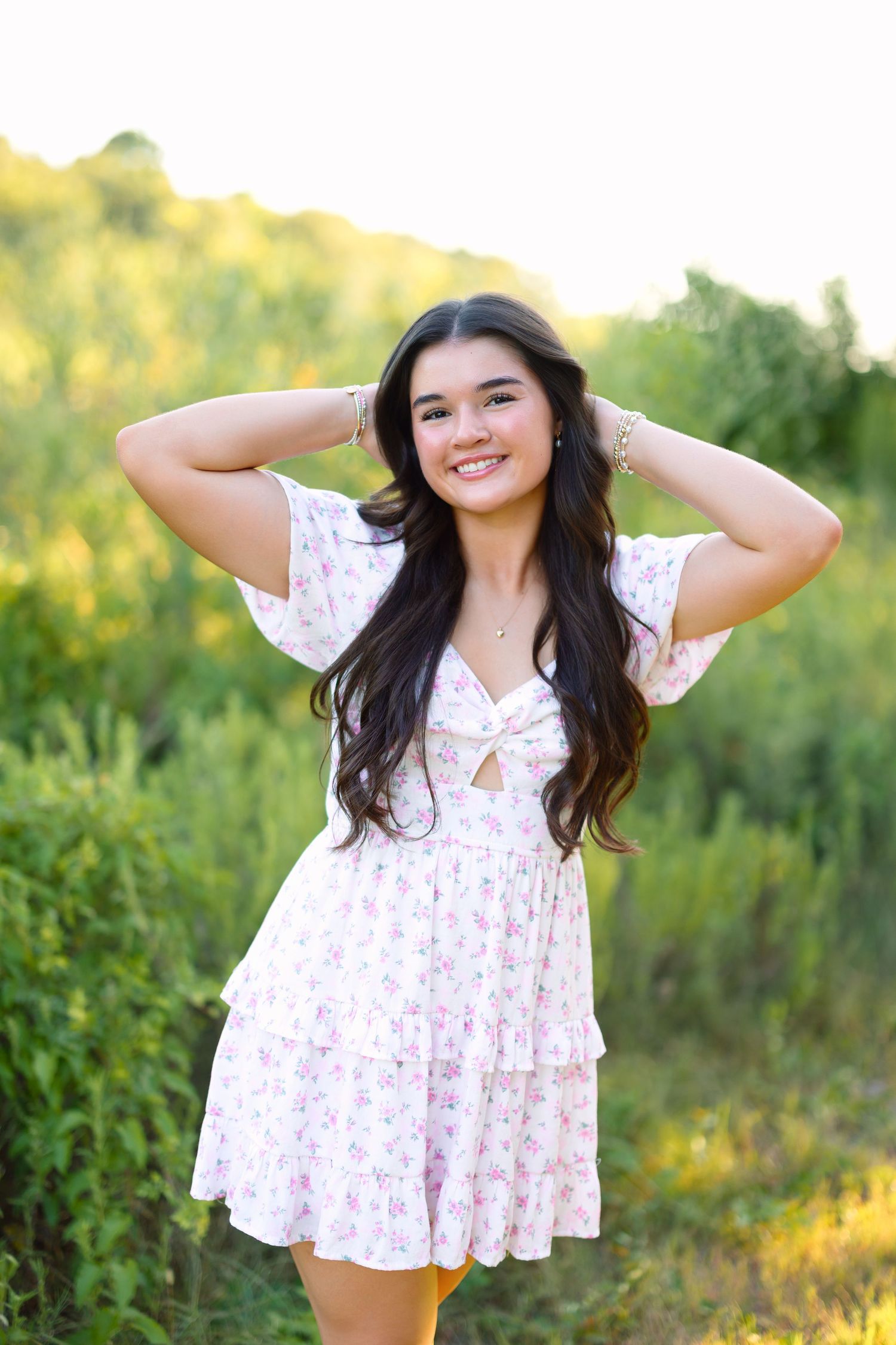 Smiling young woman in white floral dress posing outdoors with hands behind head against green foliage backdrop.