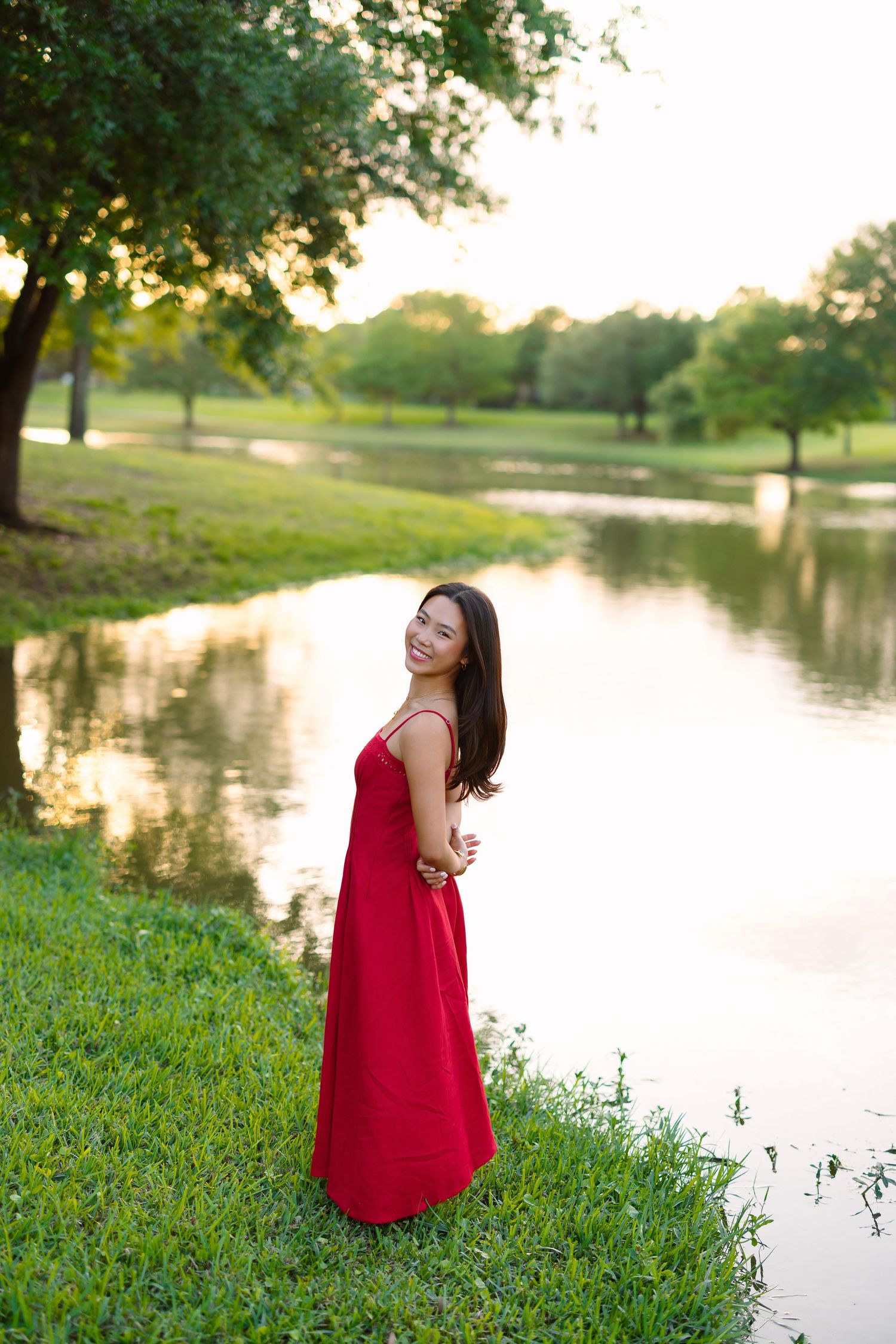 Young woman in red maxi dress smiling by a reflective pond at golden hour in a lush green park.
