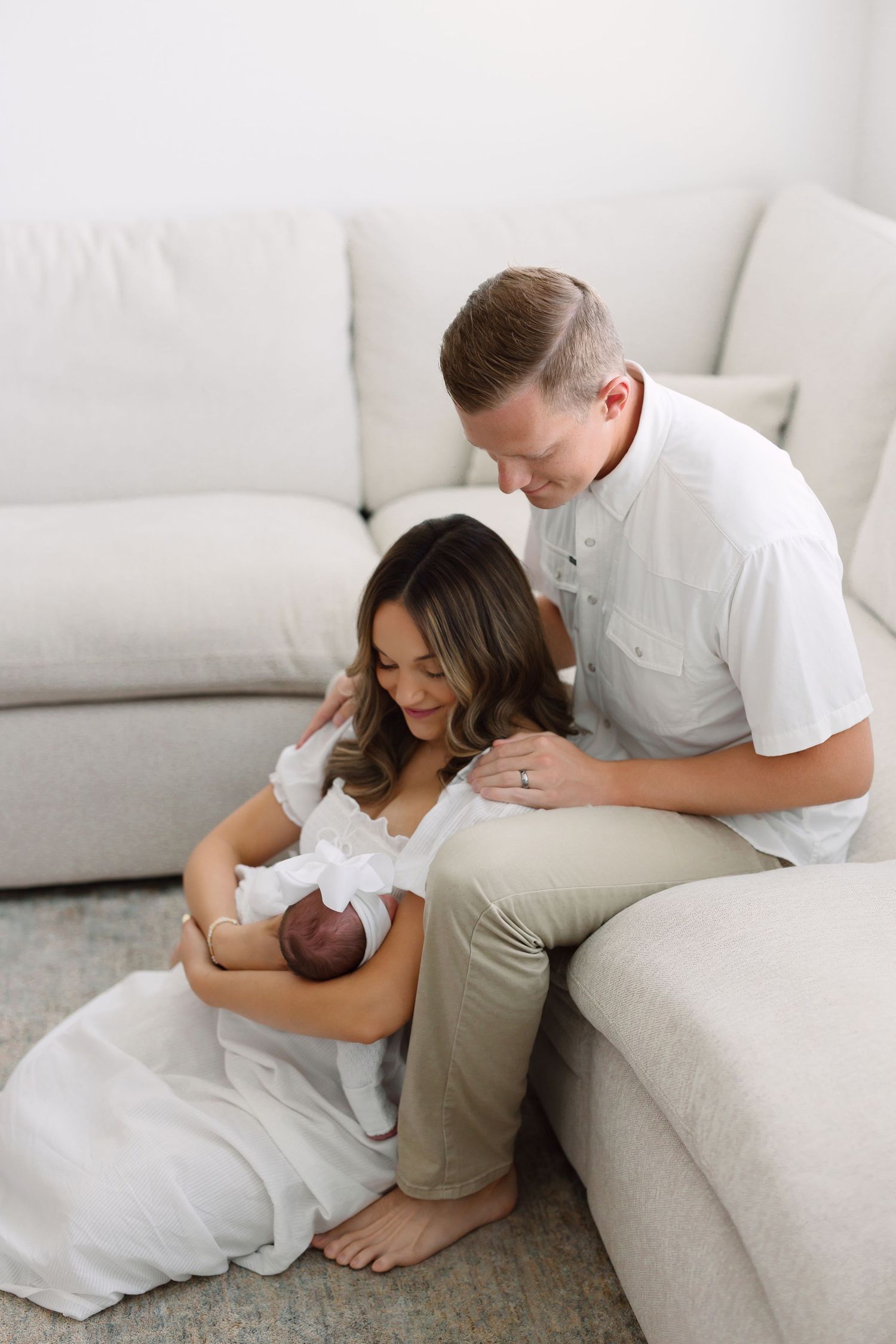 A couple sits by a white sofa, lovingly gazing at their newborn baby wrapped in white cloth.