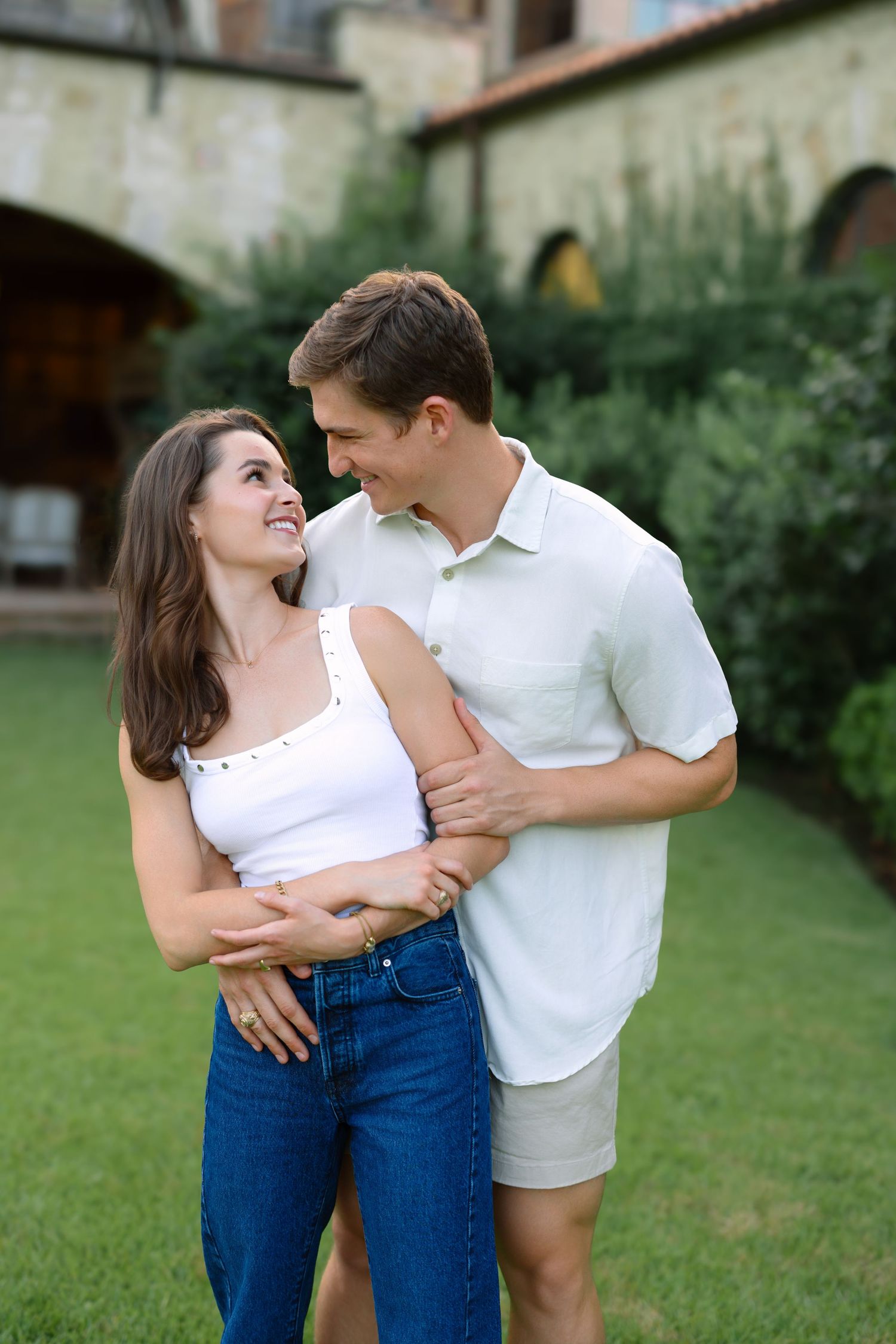 A smiling couple embraces in a lush garden courtyard, wearing white tops and jeans, gazing lovingly at each other.