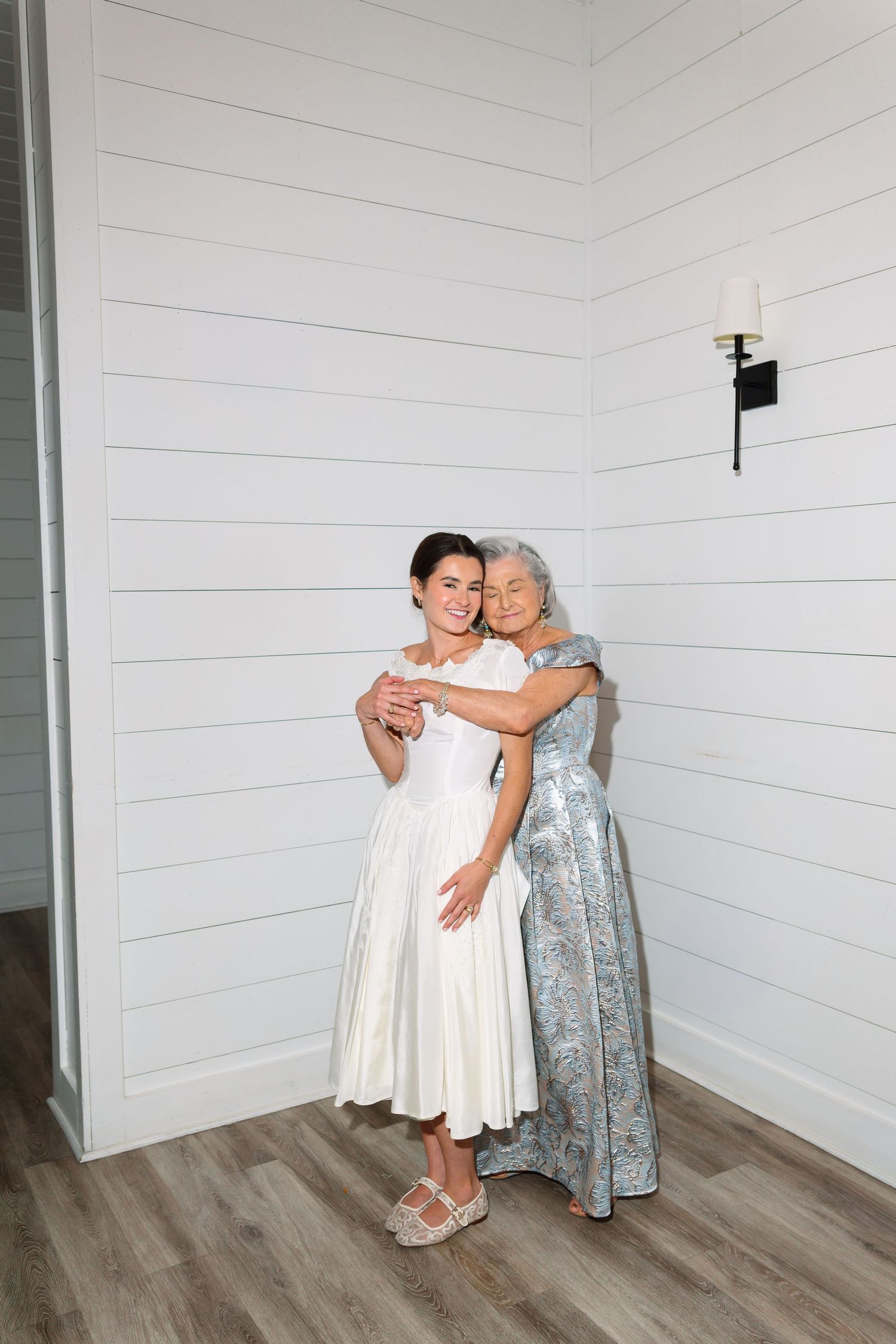 Two women in elegant dresses share a warm embrace, posing together in a bright white shiplap room.