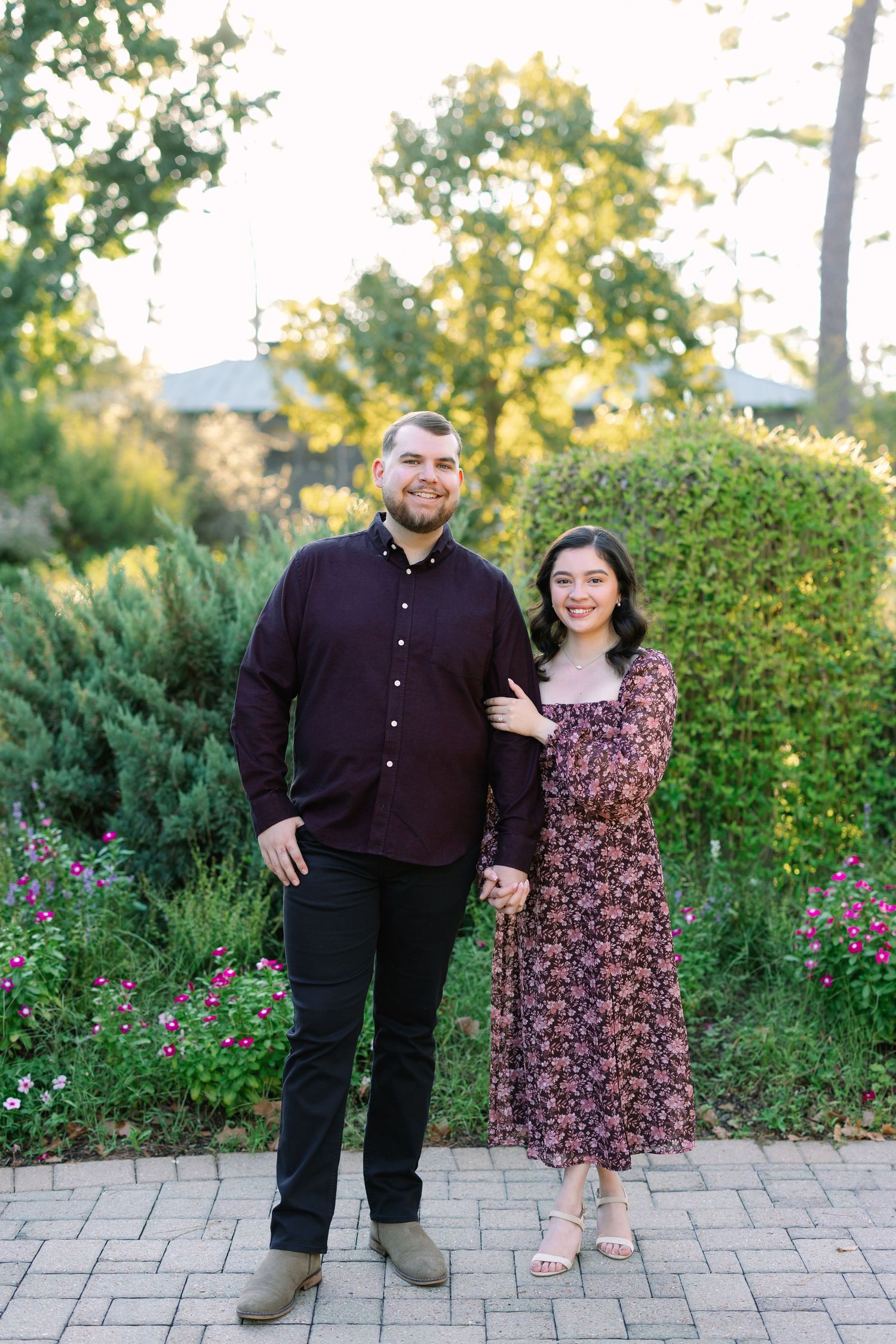 A couple in matching burgundy outfits smiling together in a lush garden with colorful flowers and greenery.