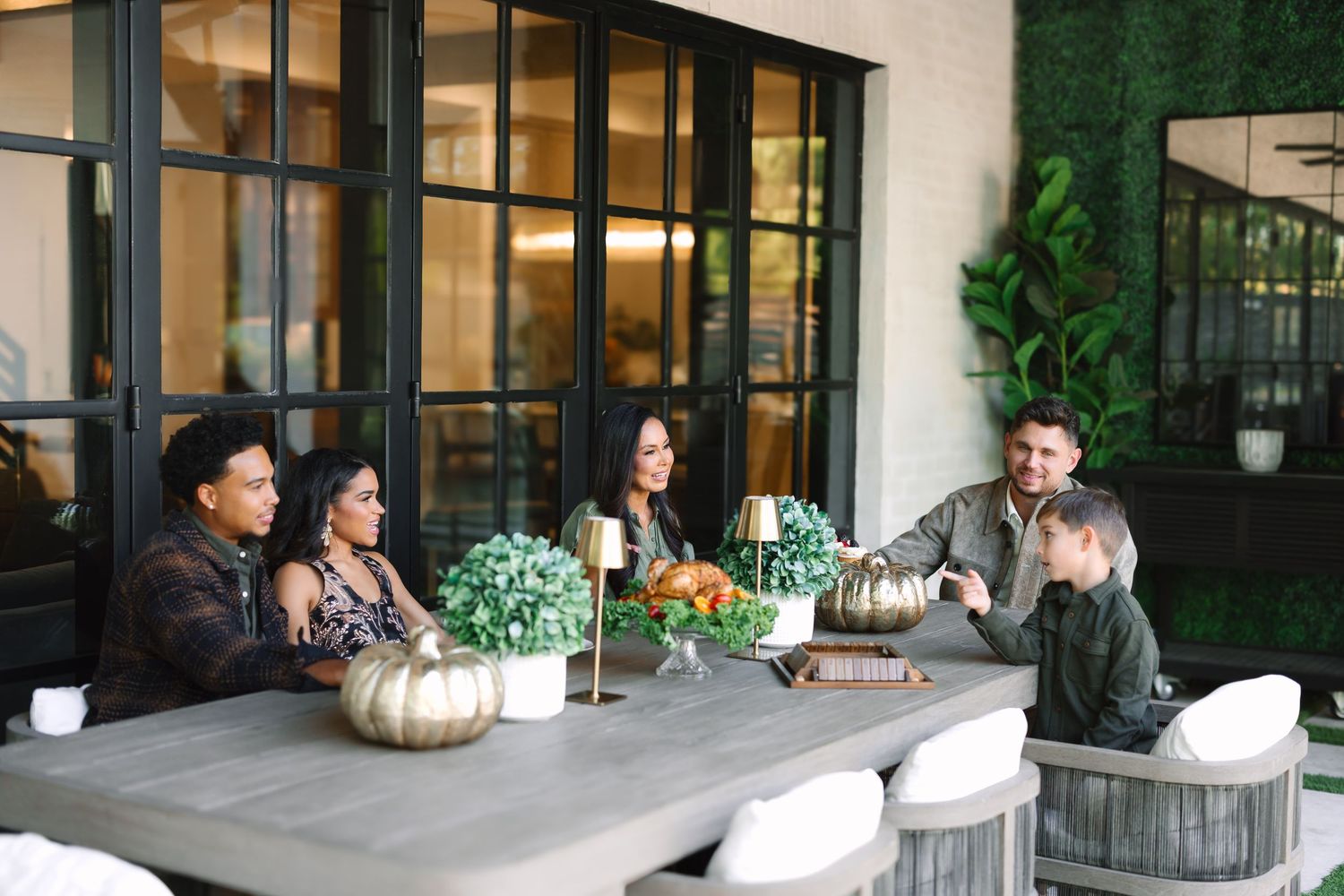 Family gathered around outdoor dining table with fall decor, pumpkins, and greenery backdrop.