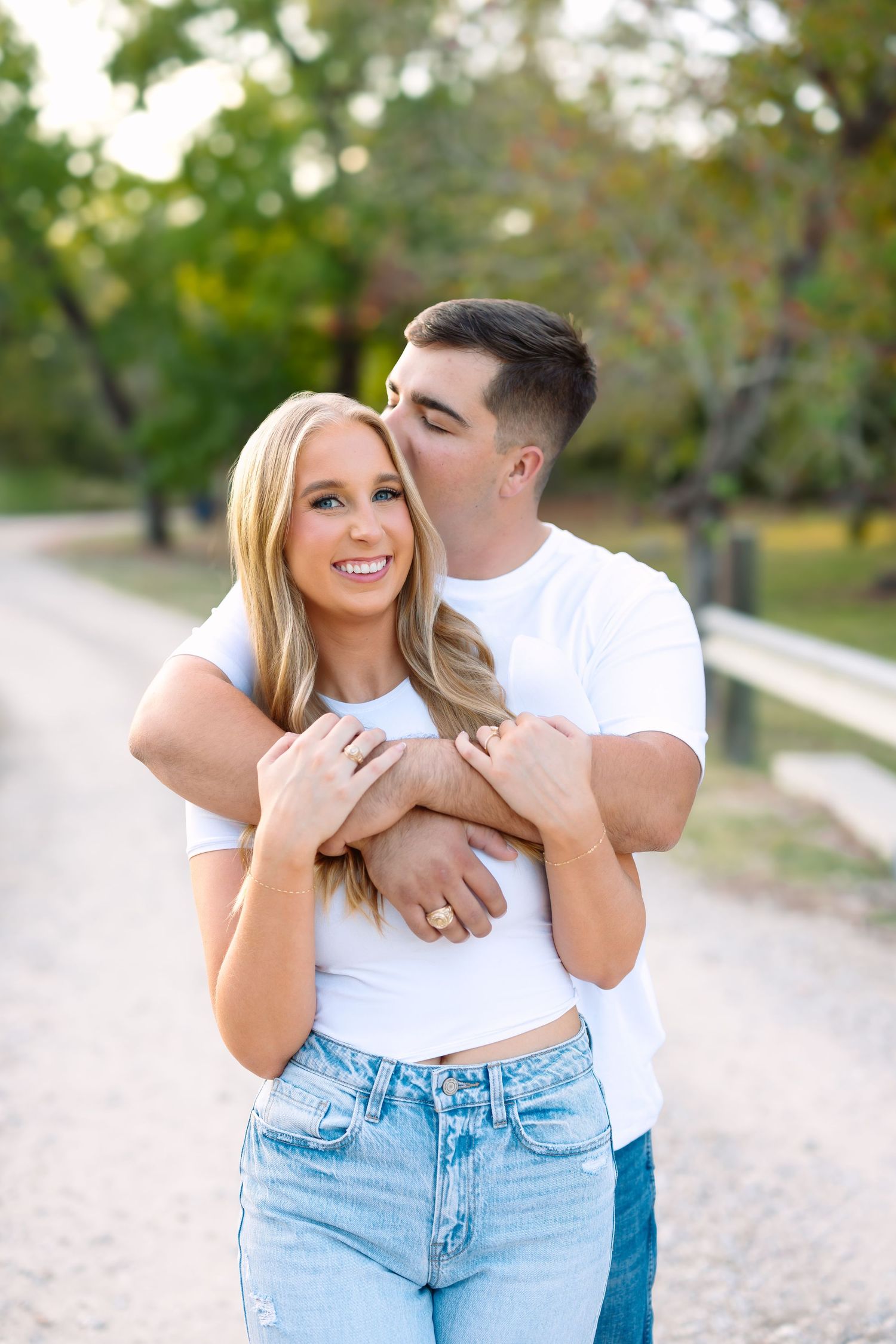 A couple in matching white tees and jeans embraces on a tree-lined path, man kissing woman's cheek.