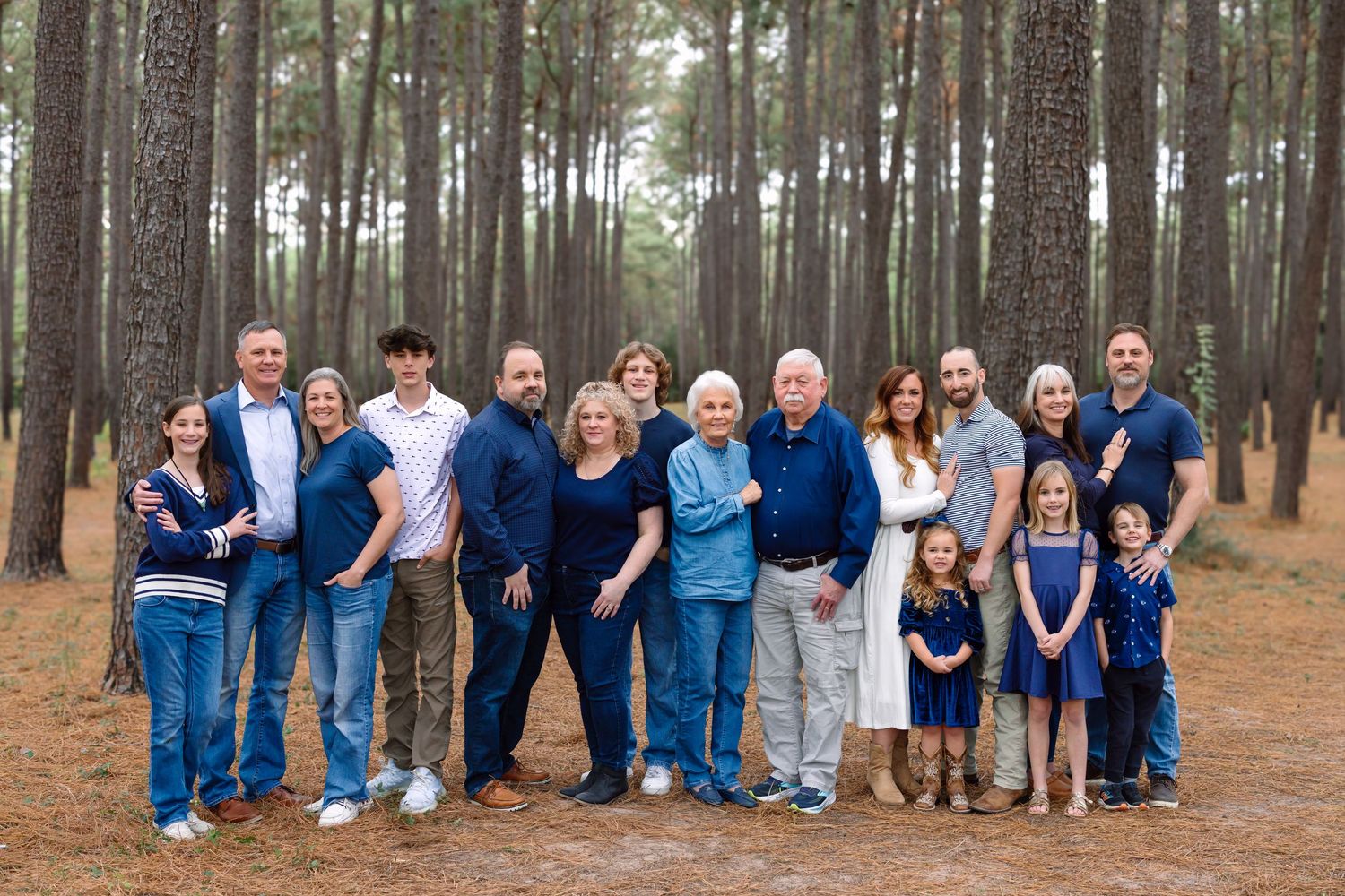 Large multigenerational family portrait outdoors among tall pine trees, dressed in coordinating navy and blue outfits.