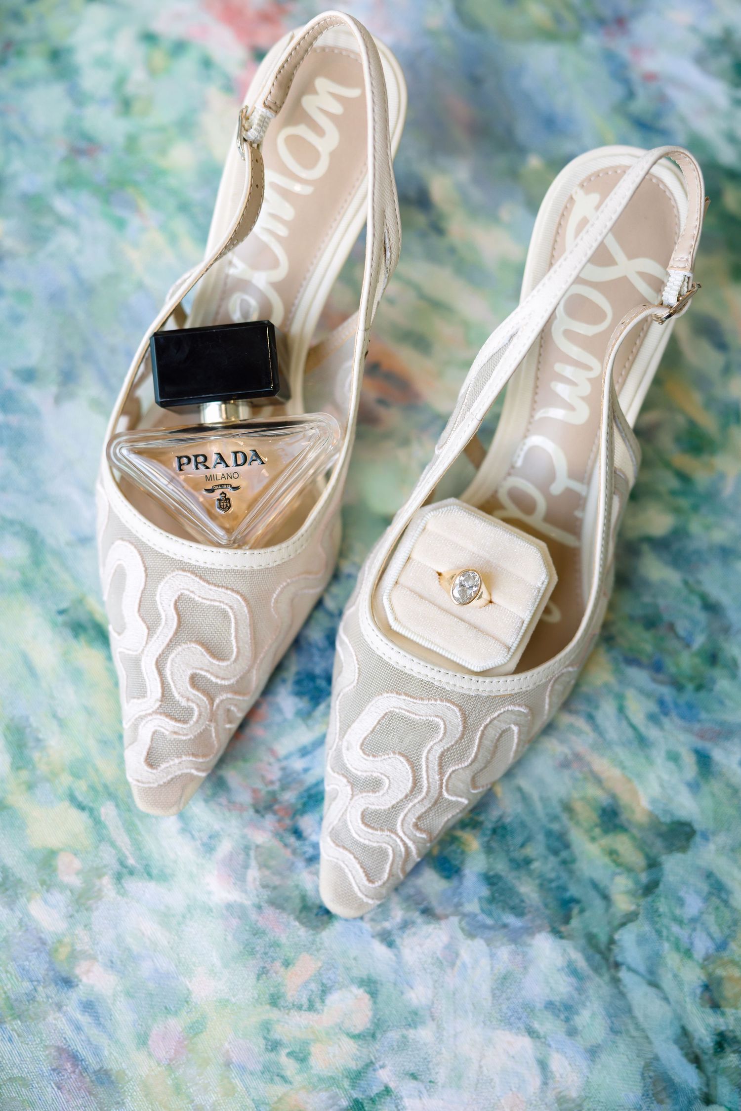 White Sam Edelman slingback heels with Prada perfume bottle and diamond engagement ring displayed on floral surface.