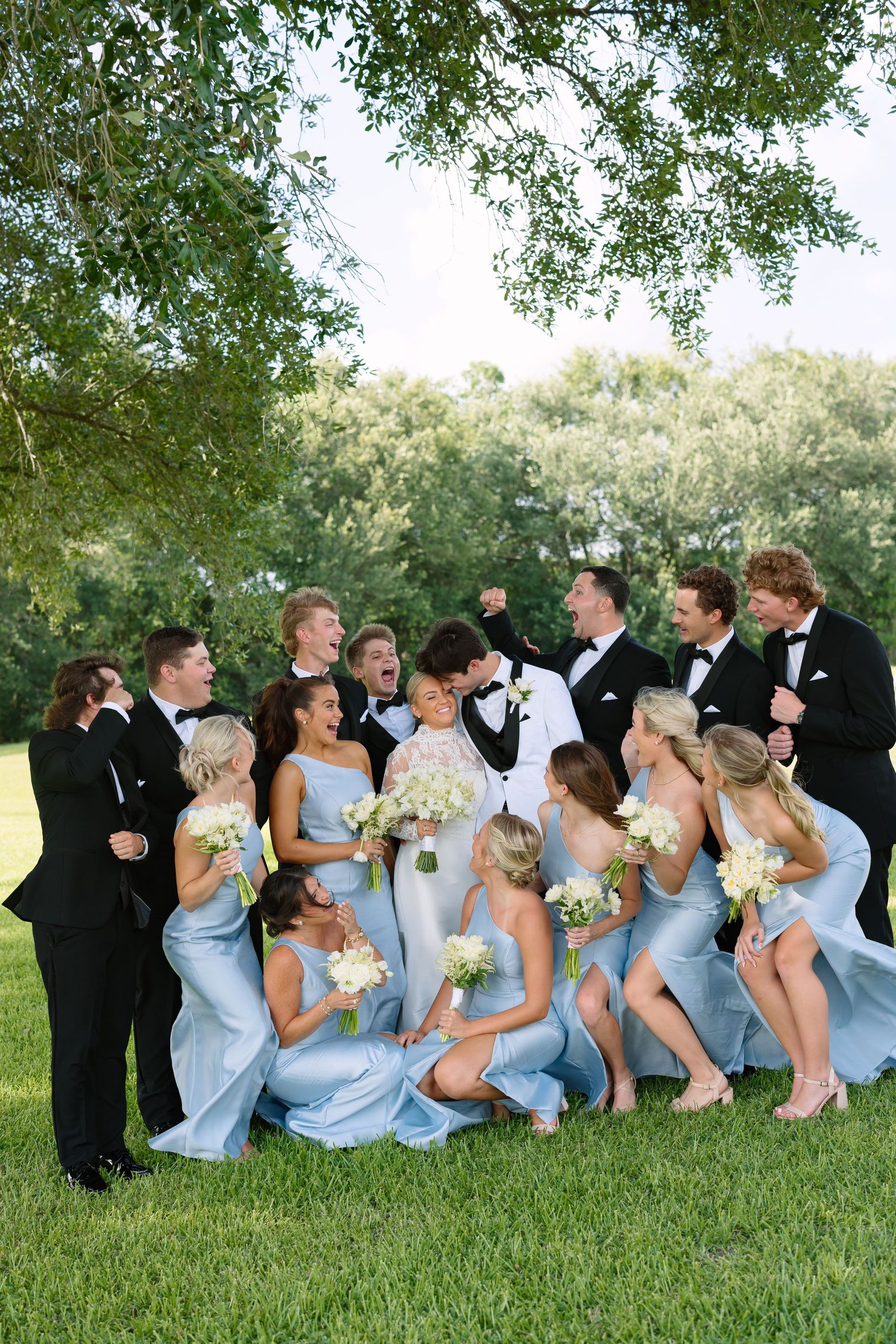 Wedding party in light blue gowns and black tuxedos celebrating outdoors under lush green trees on a sunny day.
