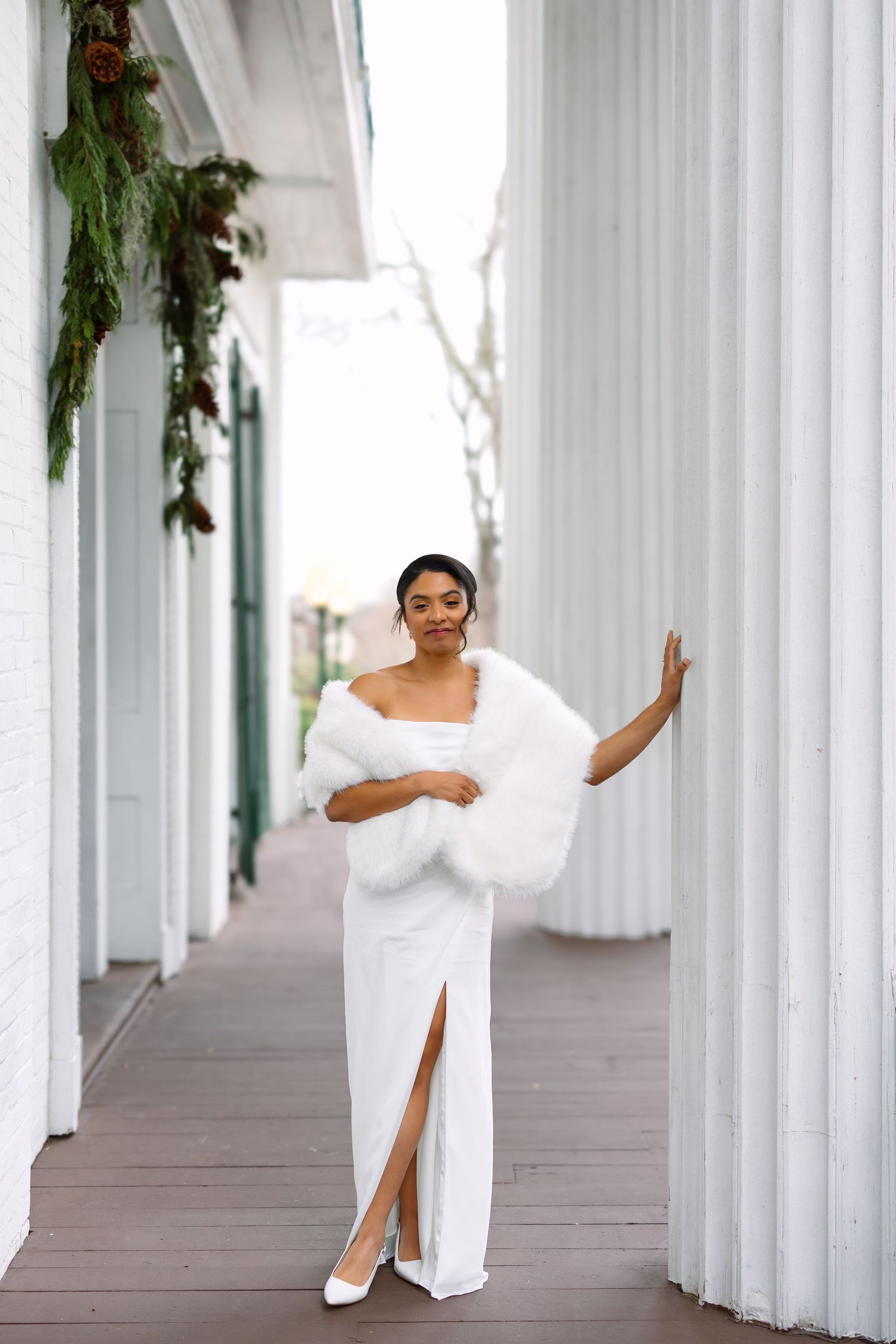 Bride in white gown and fur wrap poses between white columns at a winter wedding venue.