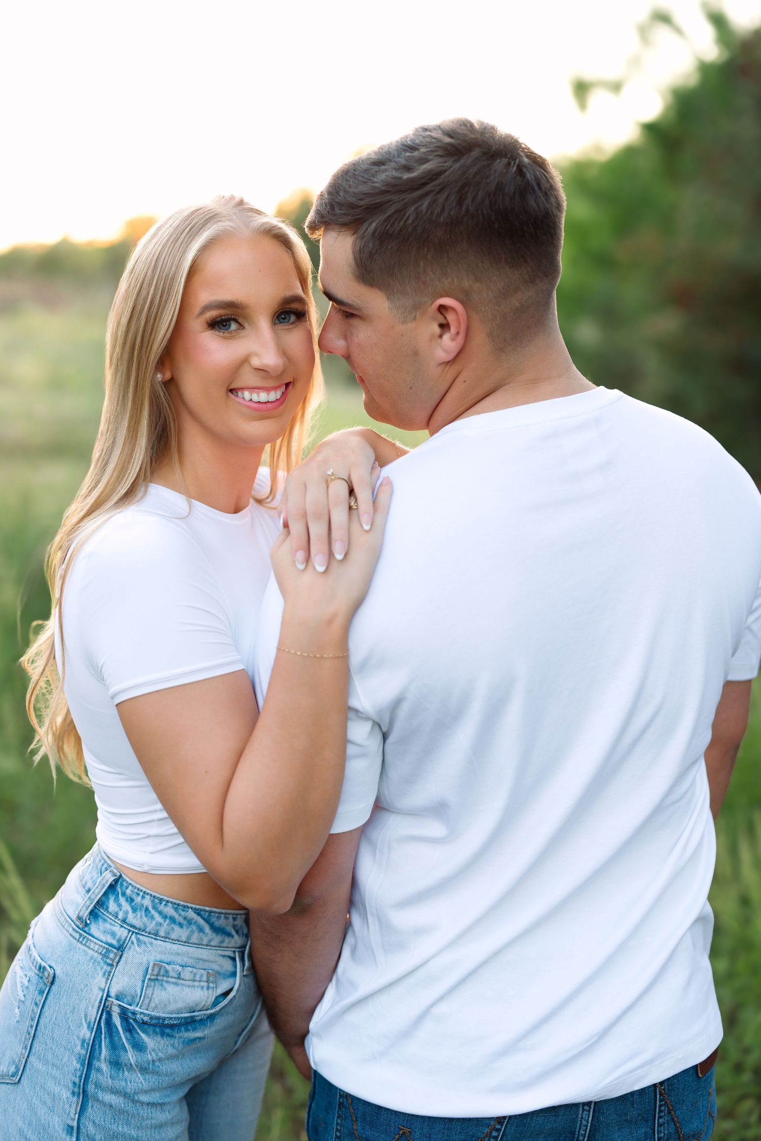 A smiling blonde woman in a white crop top embraces a man in a white t-shirt outdoors in a sunny green field.