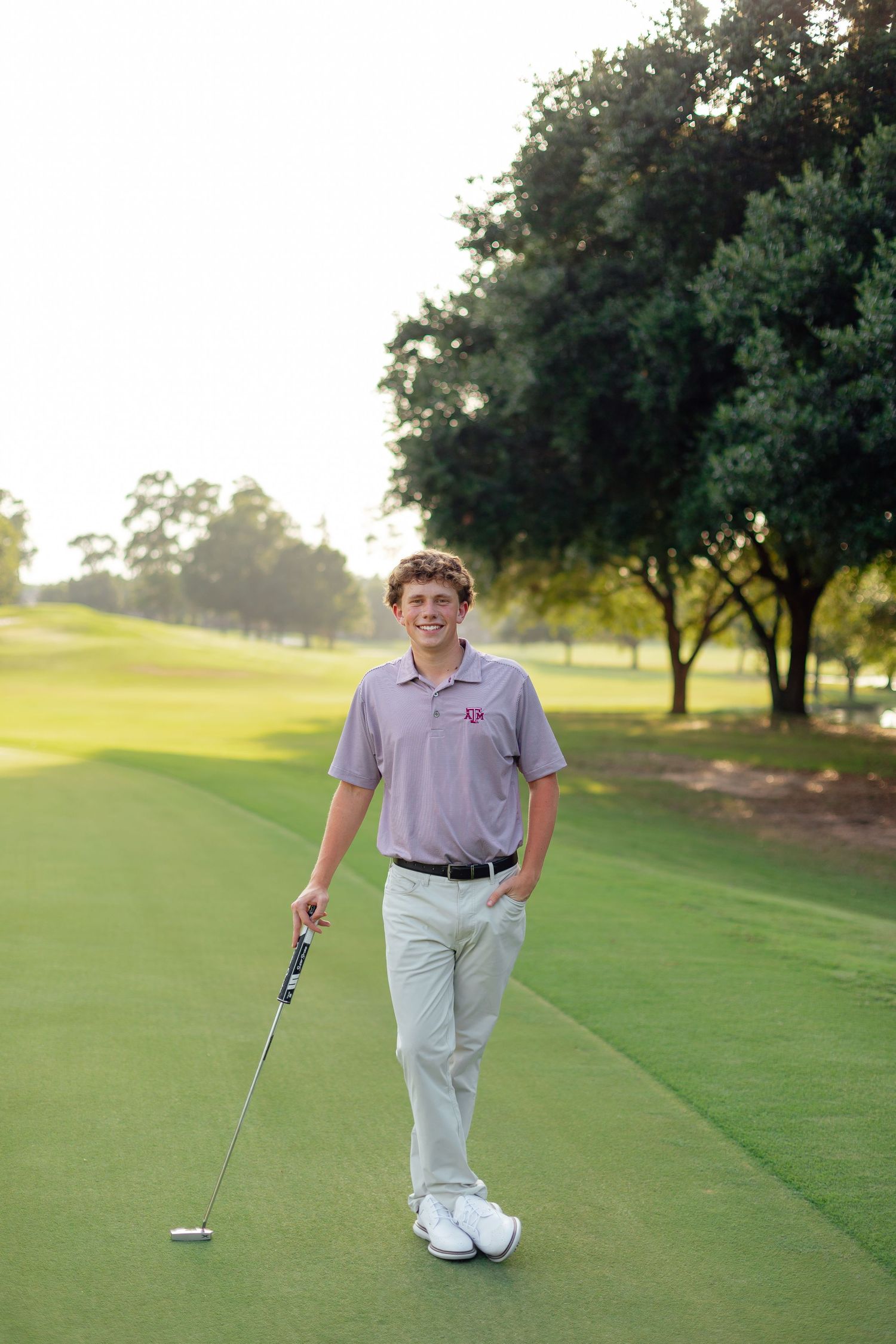 A golfer stands on a lush green fairway with trees in the background during a sunny afternoon.