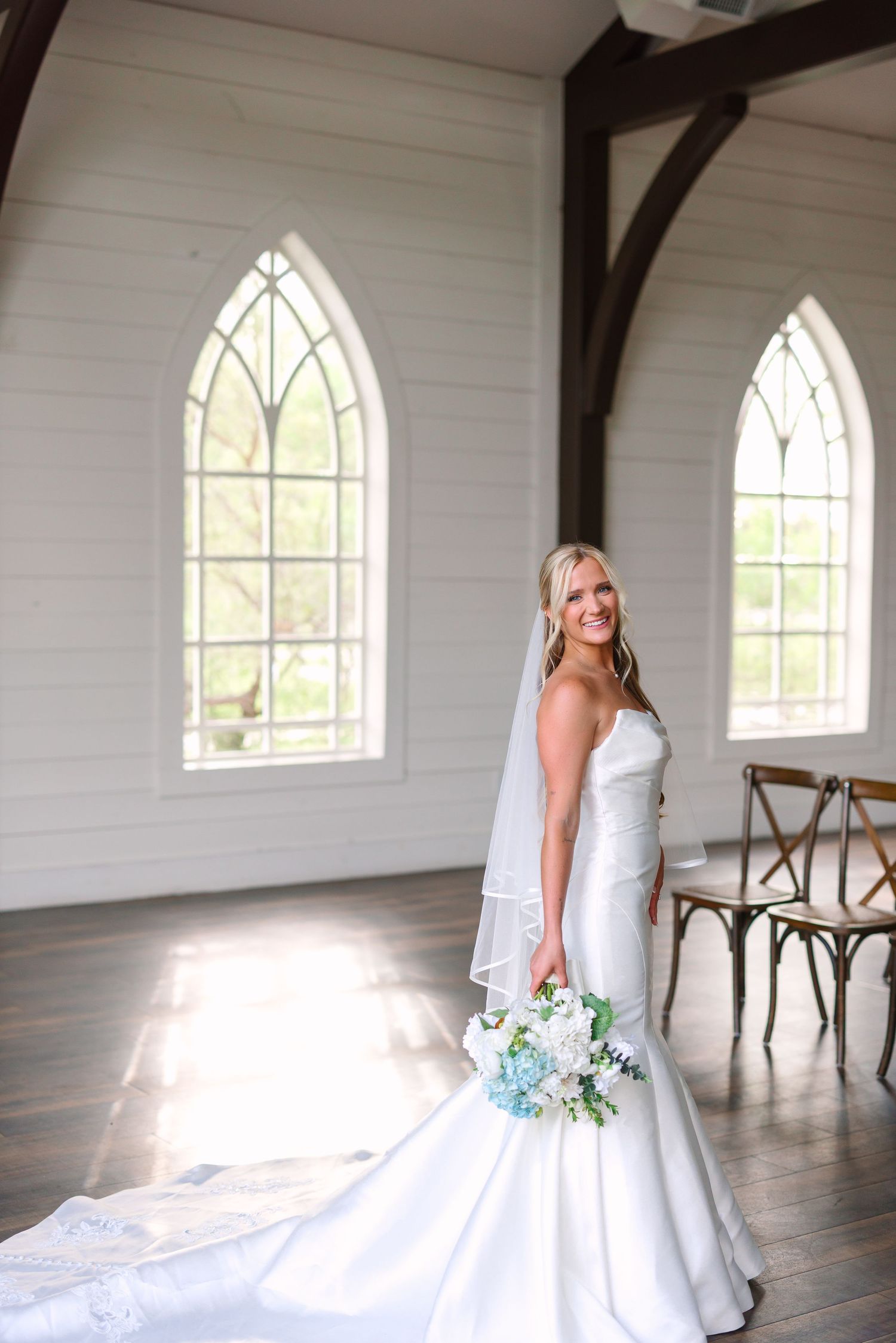 A bride in a white wedding gown poses in a chapel with arched windows and white walls.