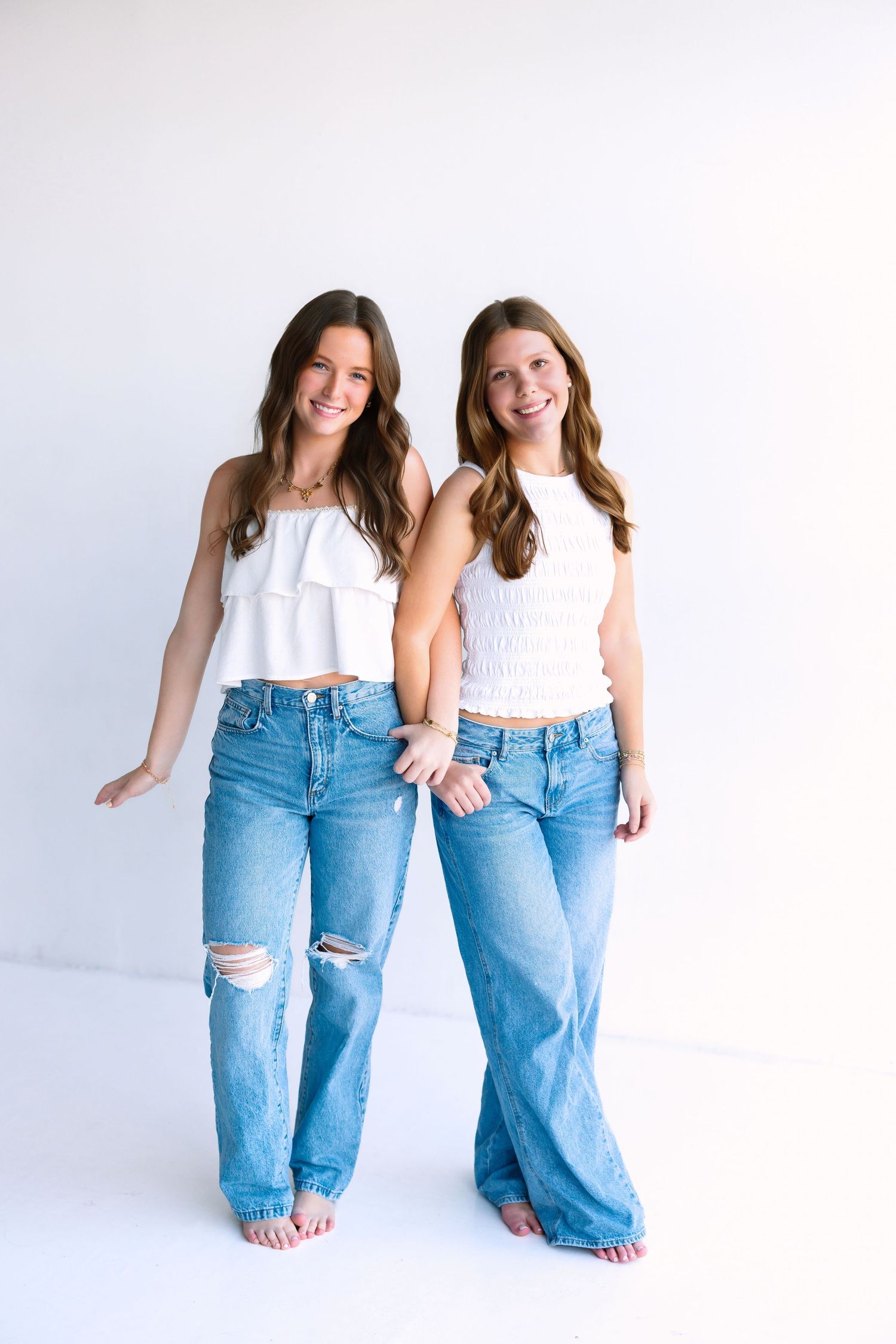 Two women in matching white tops and distressed jeans posing playfully in studio.