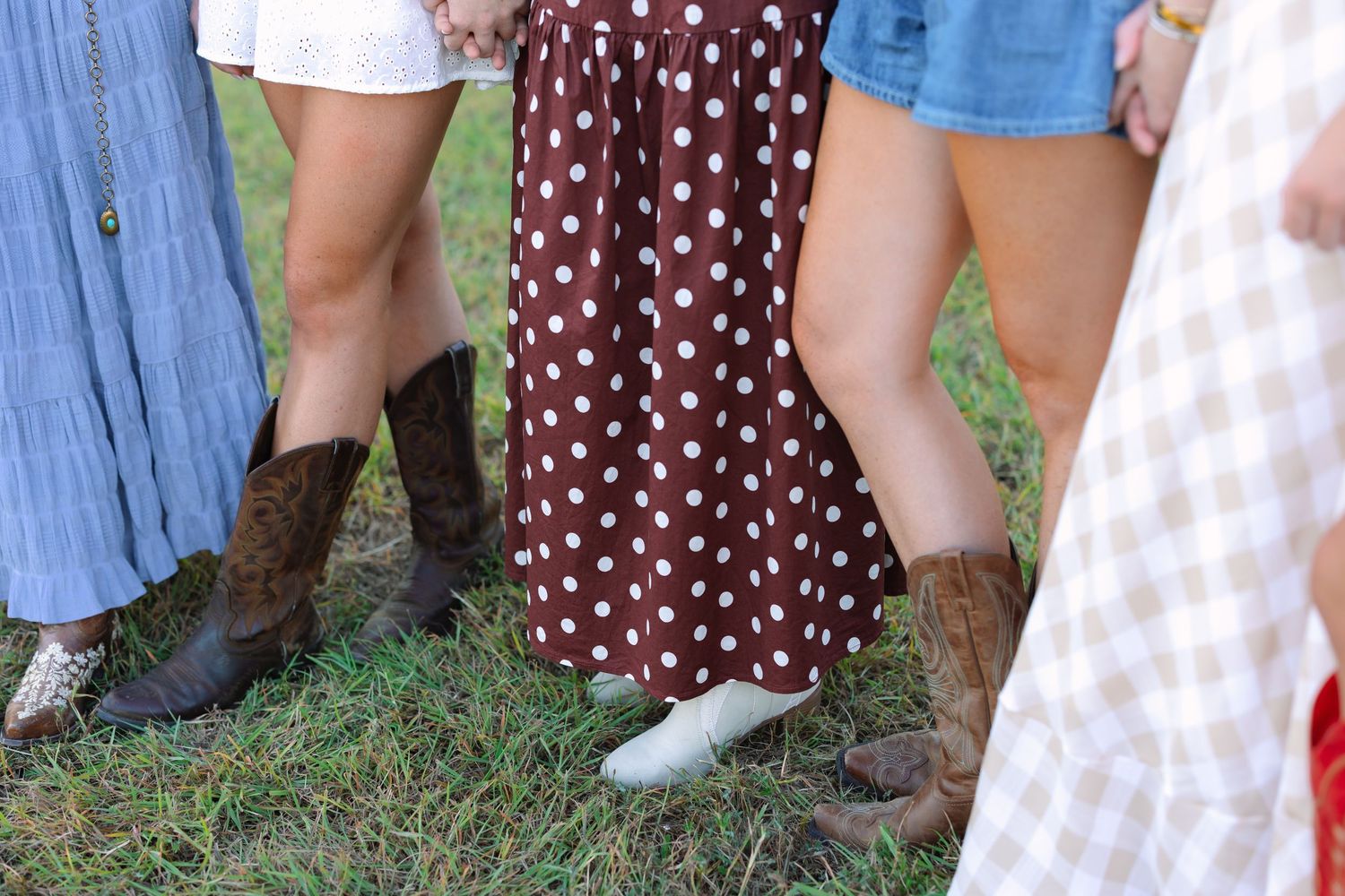 Close-up of cowboy boots and dress hems on grass in front of a barn.