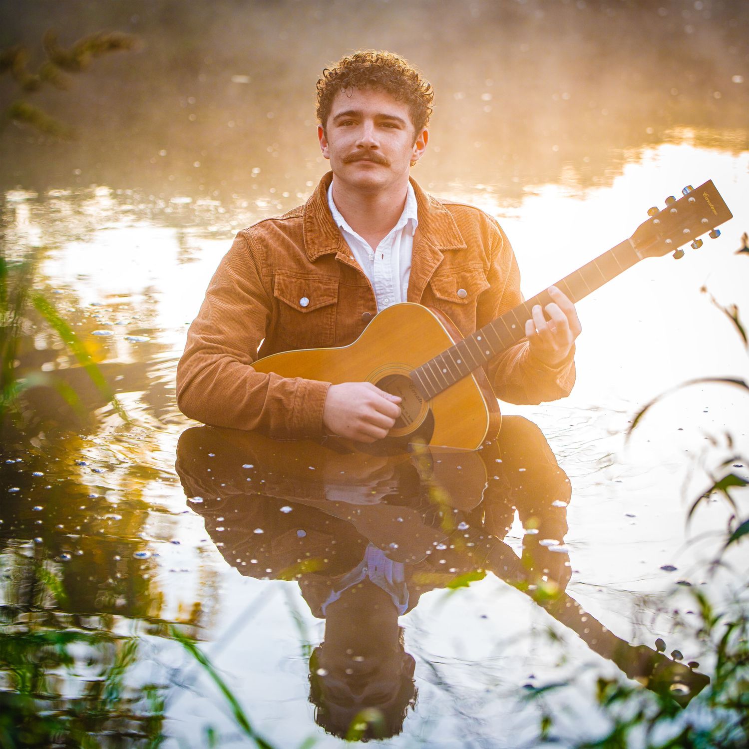 Musician playing acoustic guitar by a reflective lake at sunset creating a peaceful outdoor performance scene.