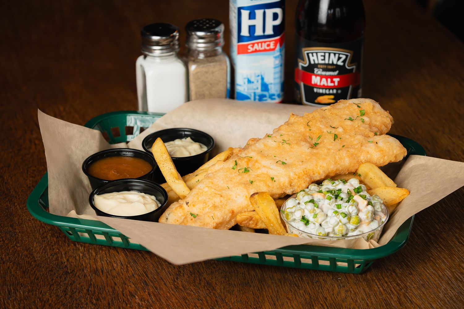Fish and chips basket with coleslaw and dipping sauce served on green tray liner.