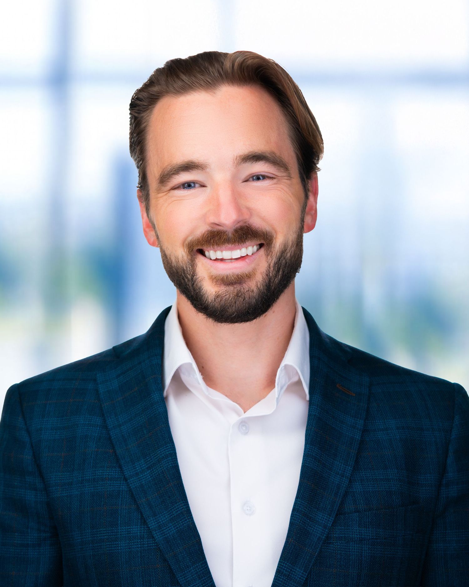 Professional headshot of person in navy suit and white shirt smiling against blurred office background.