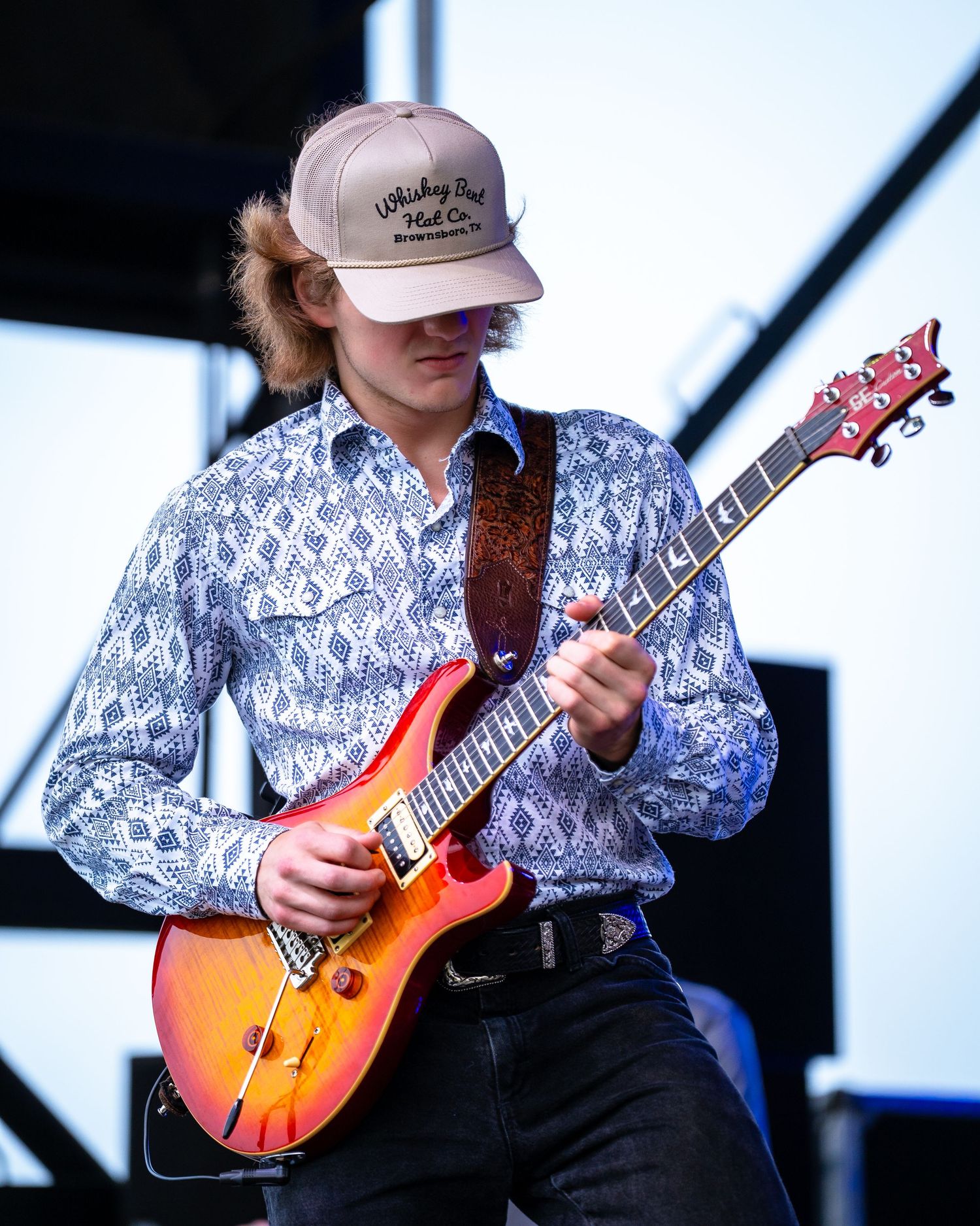 Musician wearing patterned shirt and baseball cap playing sunburst electric guitar on stage.