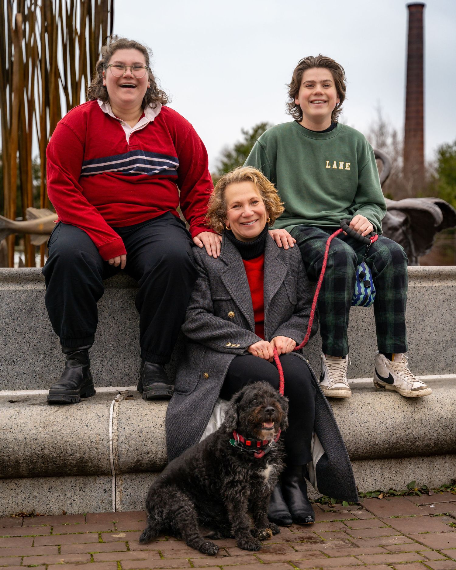 Three people sitting on stone steps outdoors with a small black dog, wearing casual winter clothing.