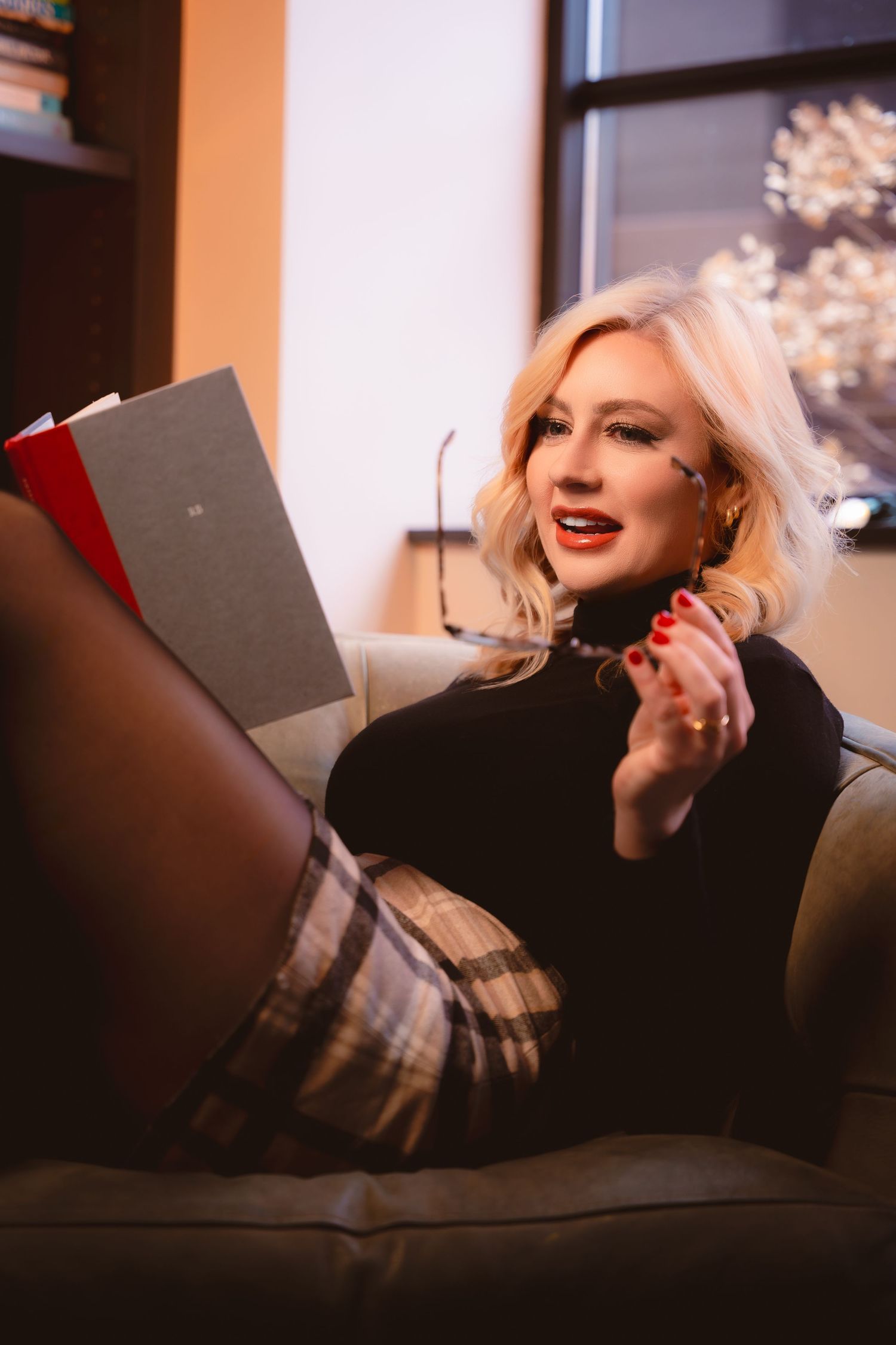 A person with platinum blonde hair wearing a black turtleneck relaxes on a couch while reading a book.