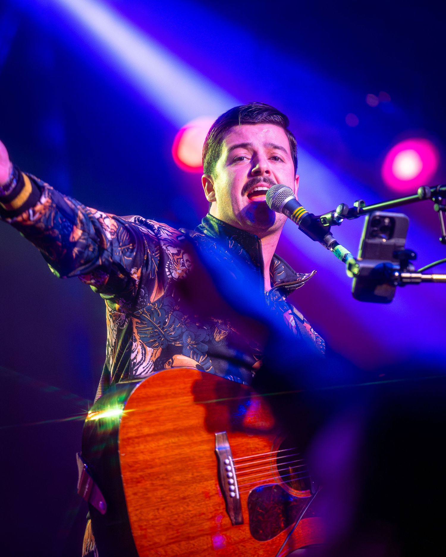 Performer with acoustic guitar singing under dramatic blue spotlights on stage.